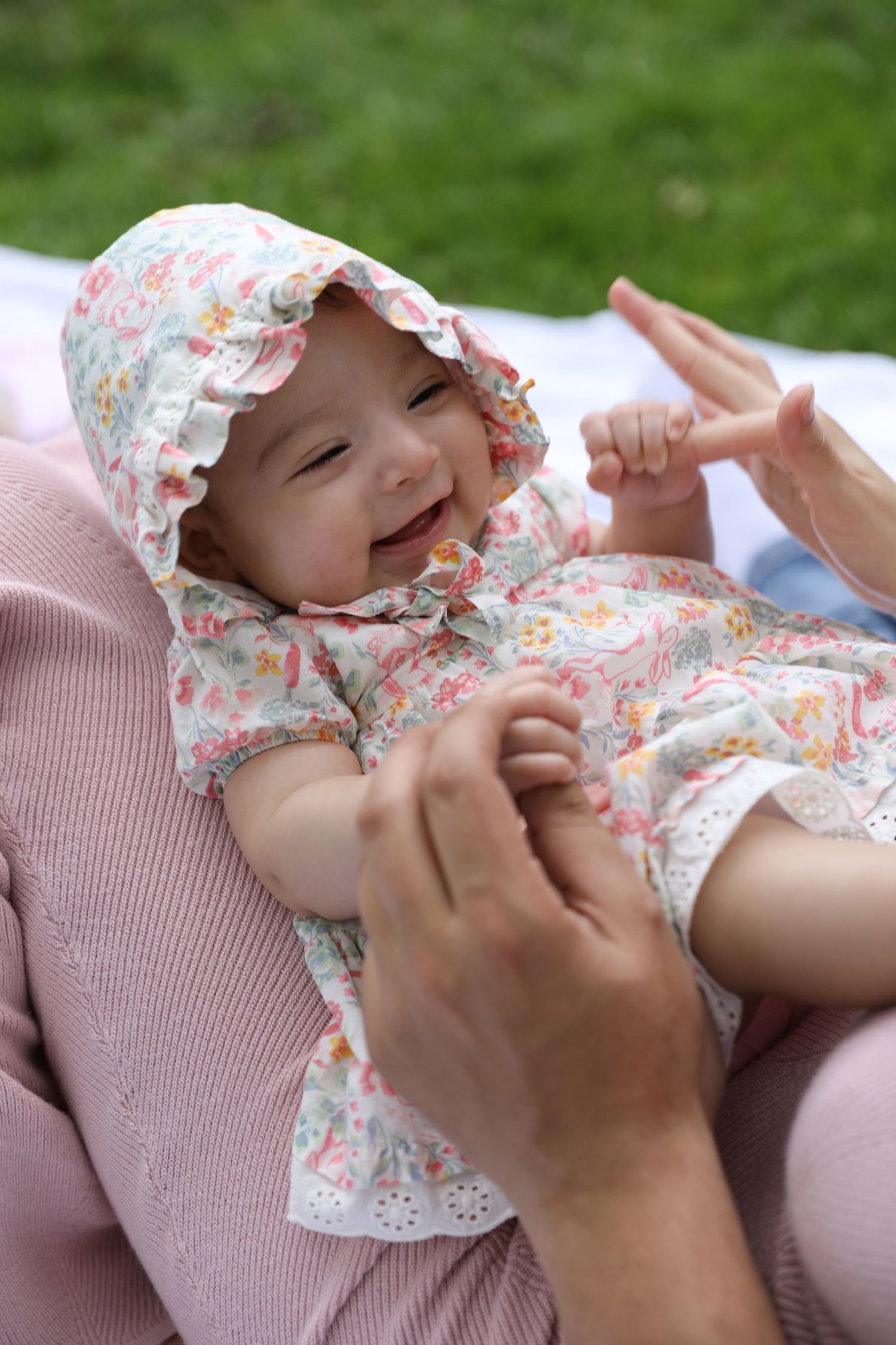 A baby wearing a floral dress and bonnet laughs while being tickled on a pink blanket outdoors.