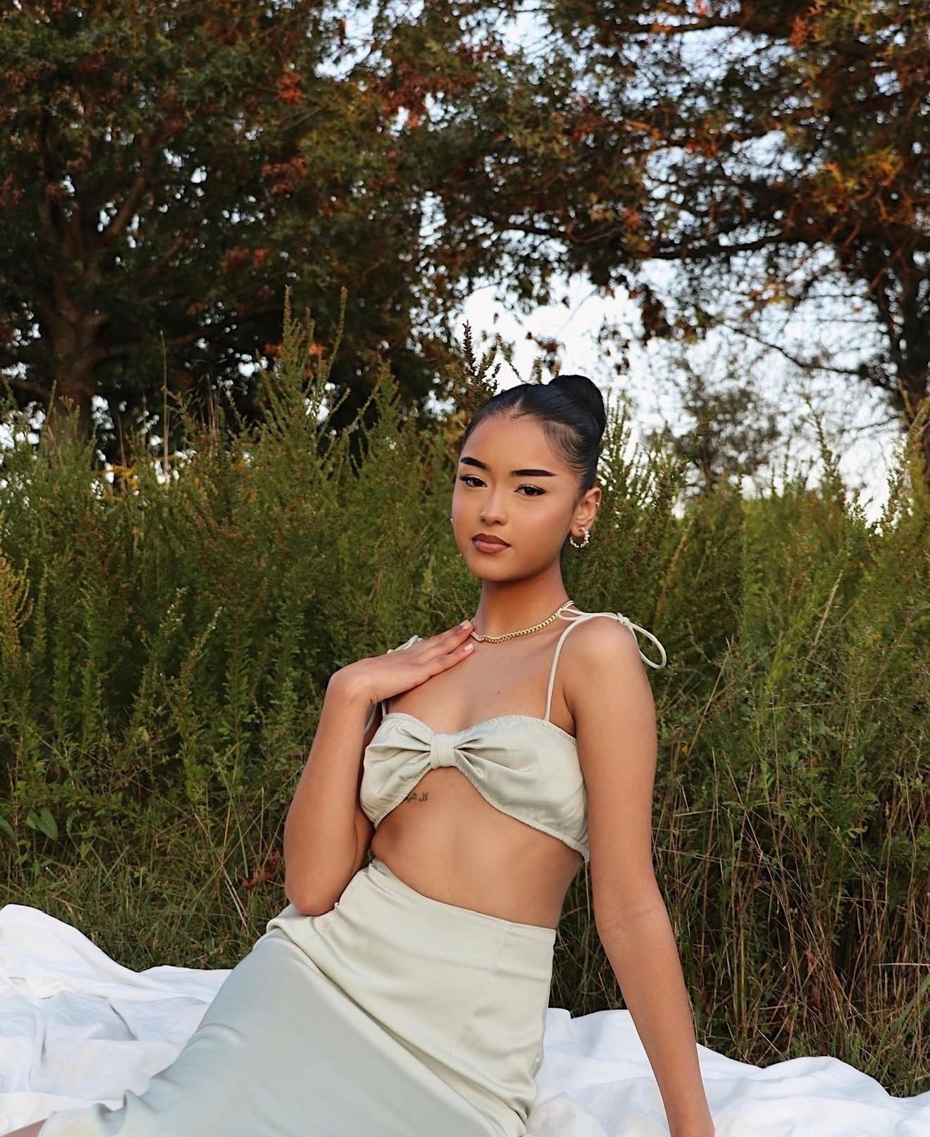 Woman in silver satin dress poses gracefully on white fabric in an outdoor garden setting during golden hour.