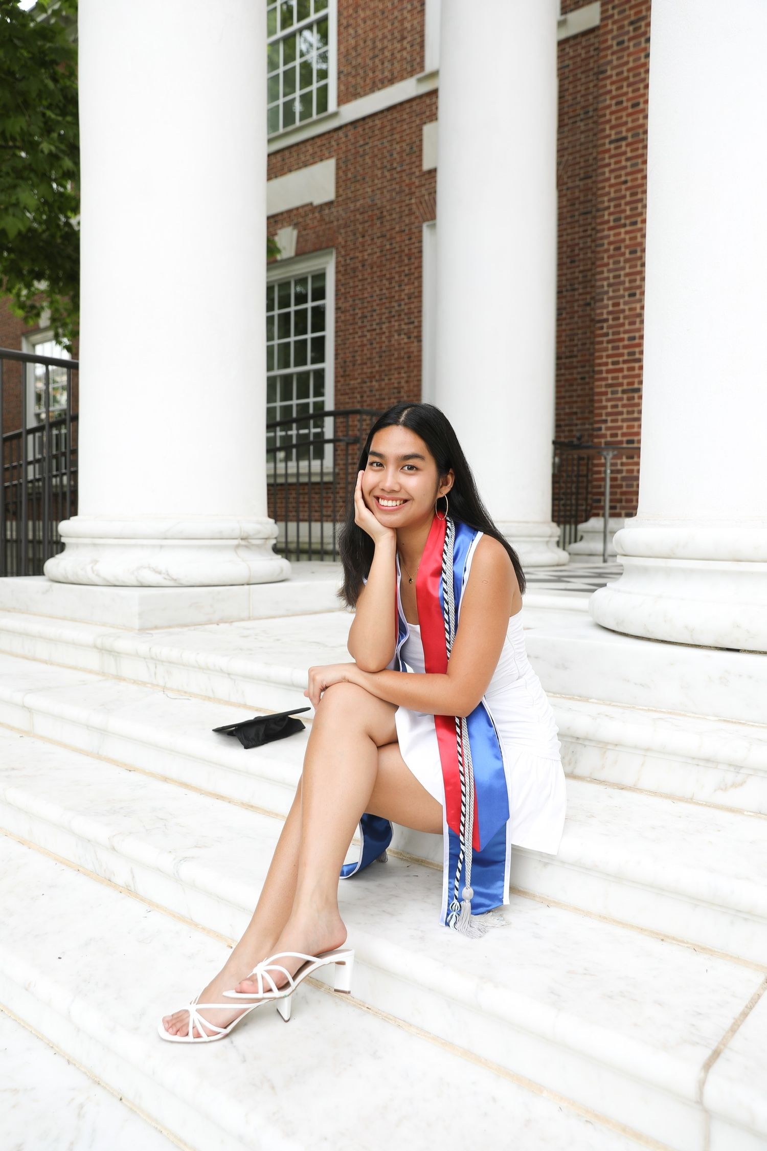 A person in a red and blue dress sits on white stone steps in front of a brick building with columns.