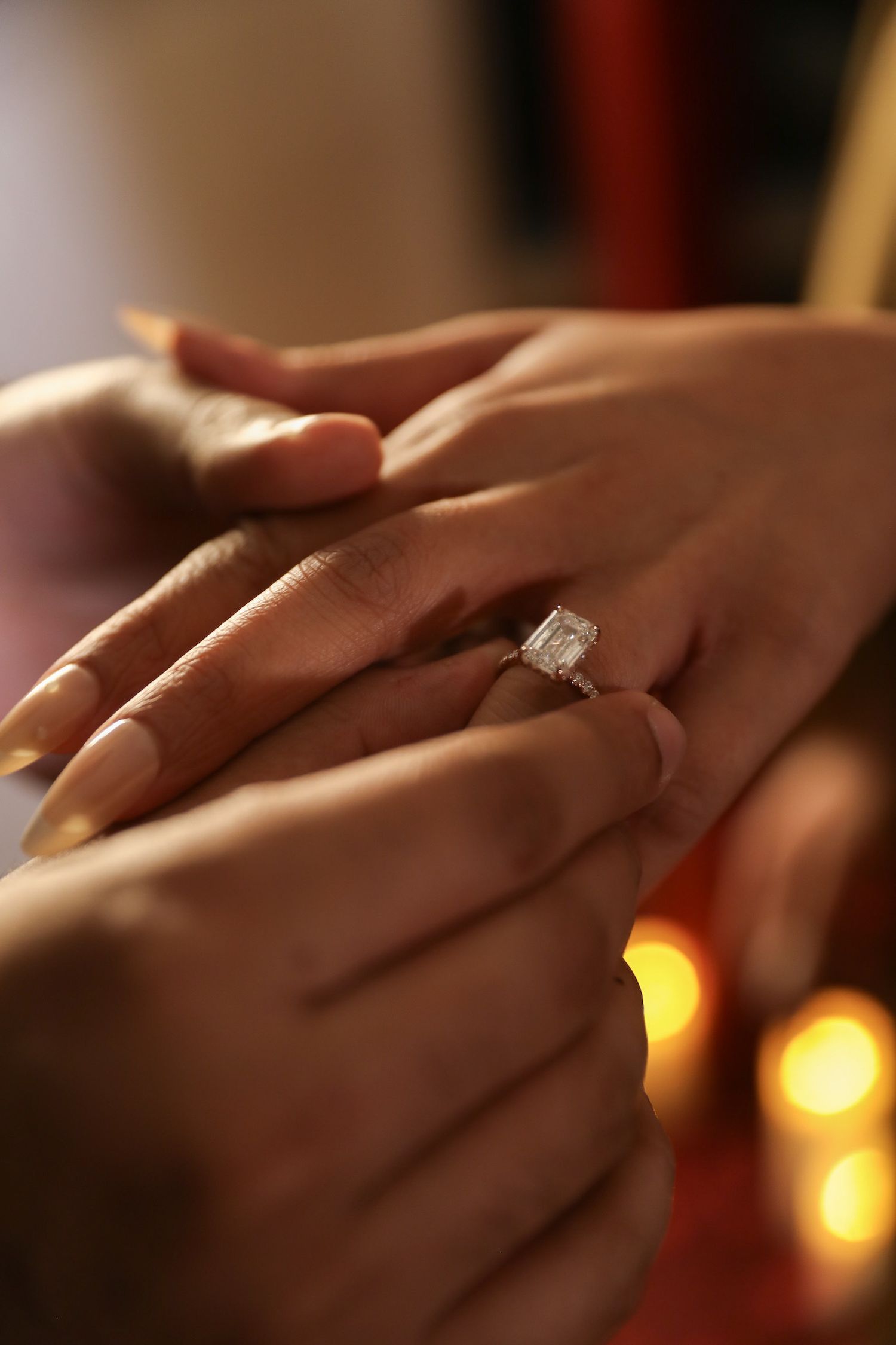 Close-up view of hands wearing a sparkling diamond engagement ring with warm glowing lights in the background.
