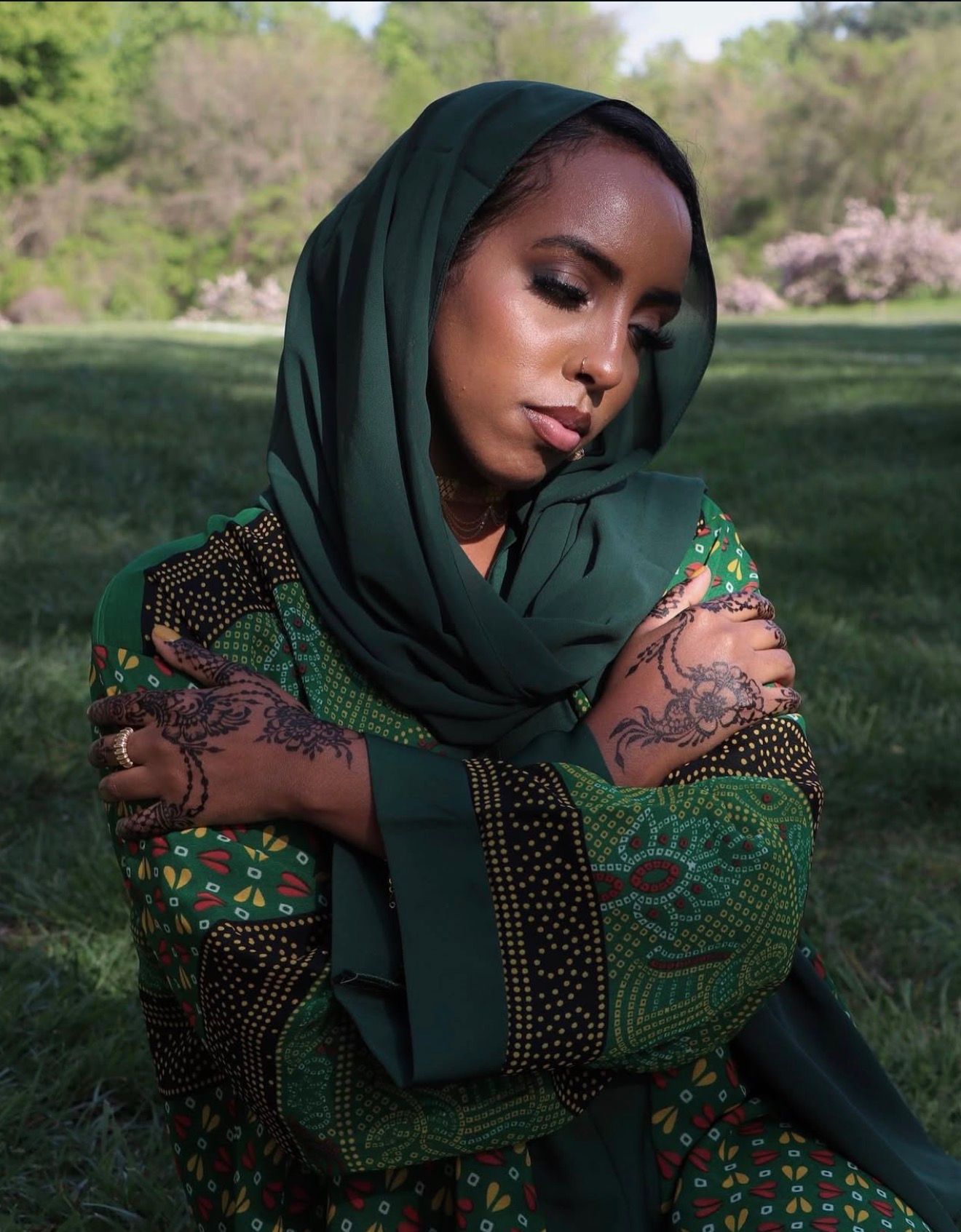 A figure in a dark hijab and patterned dress poses gracefully against a natural outdoor backdrop.