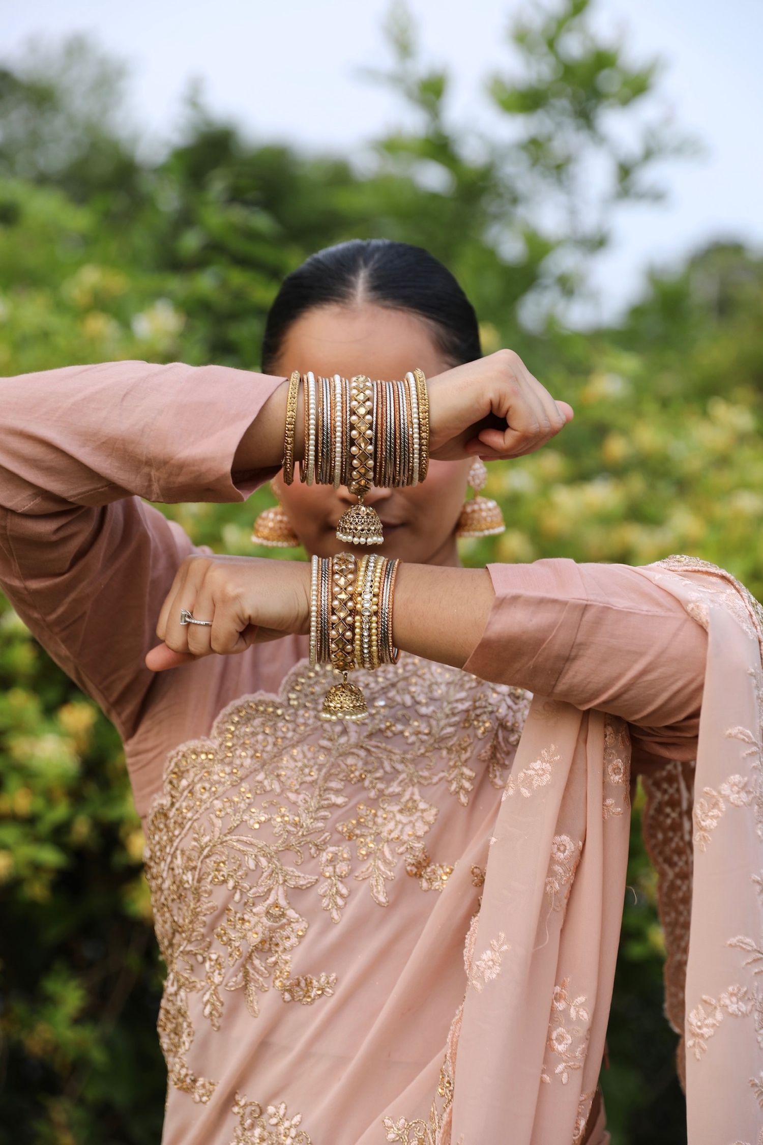 Close-up of hands adorned with gold bangles and rings wearing a blush pink embroidered dress against a garden backdrop.