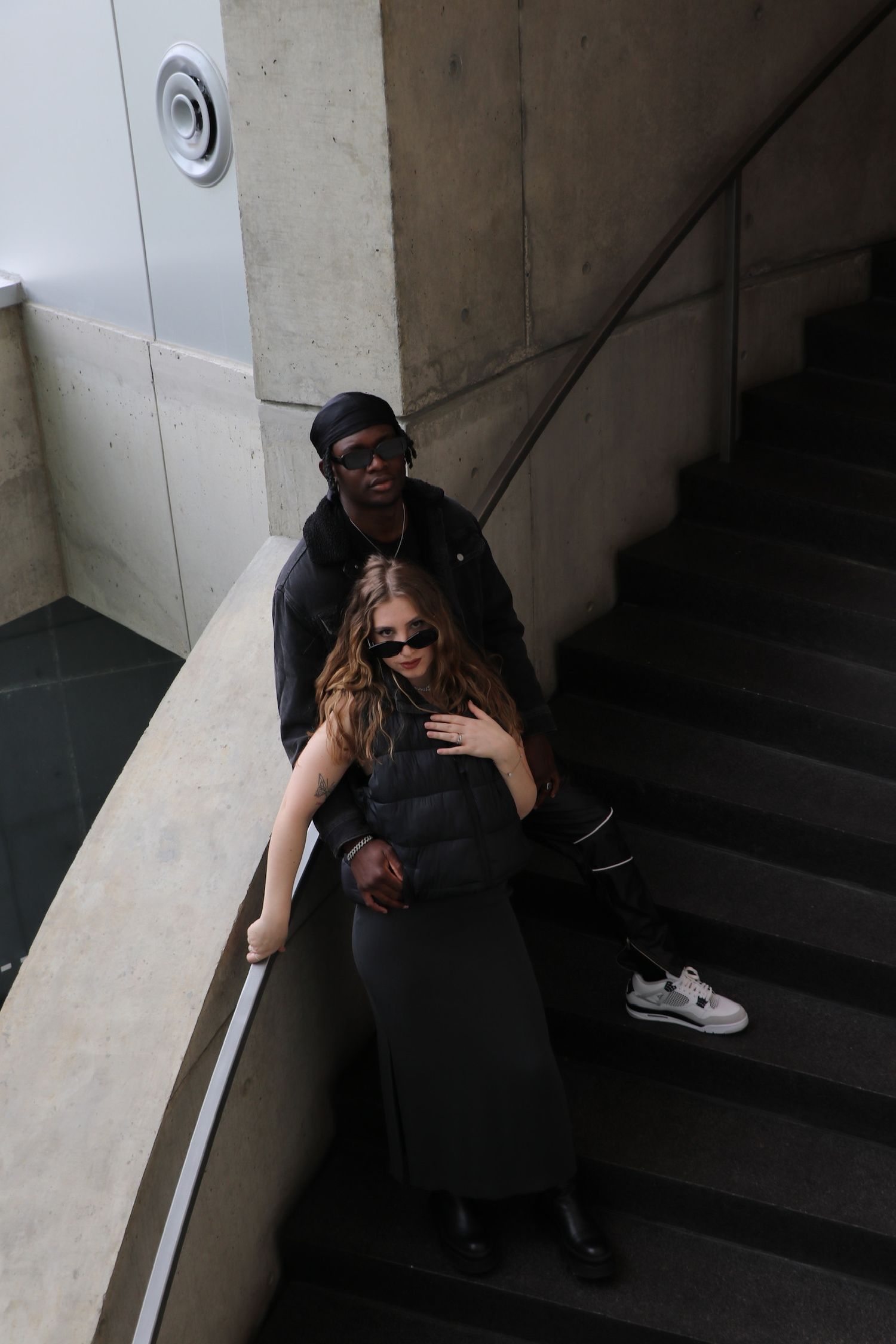 Two people in dark clothing sit together on concrete steps in a dimly lit space.