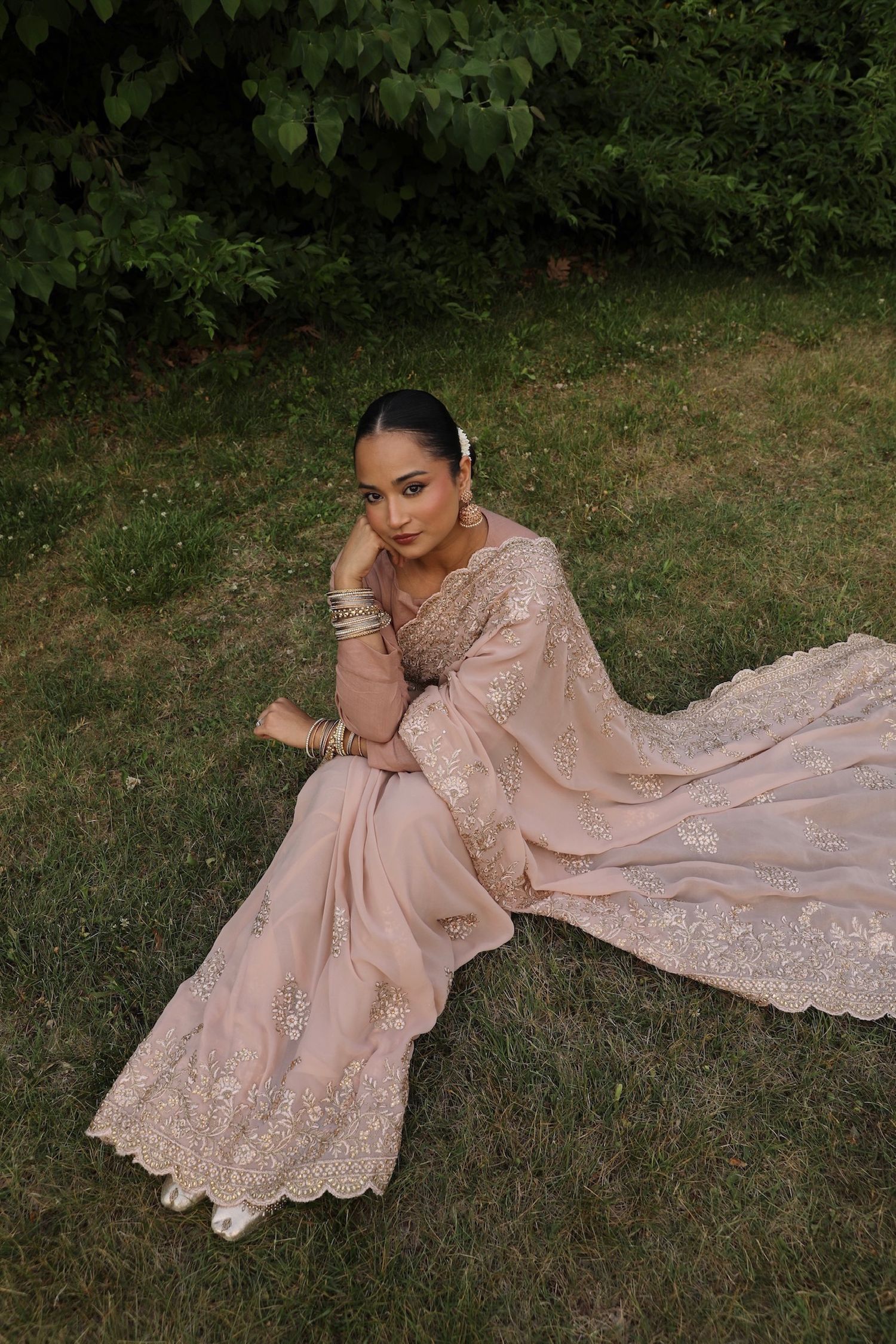 A woman in an elegant blush pink embroidered gown poses gracefully on grass in a garden setting.