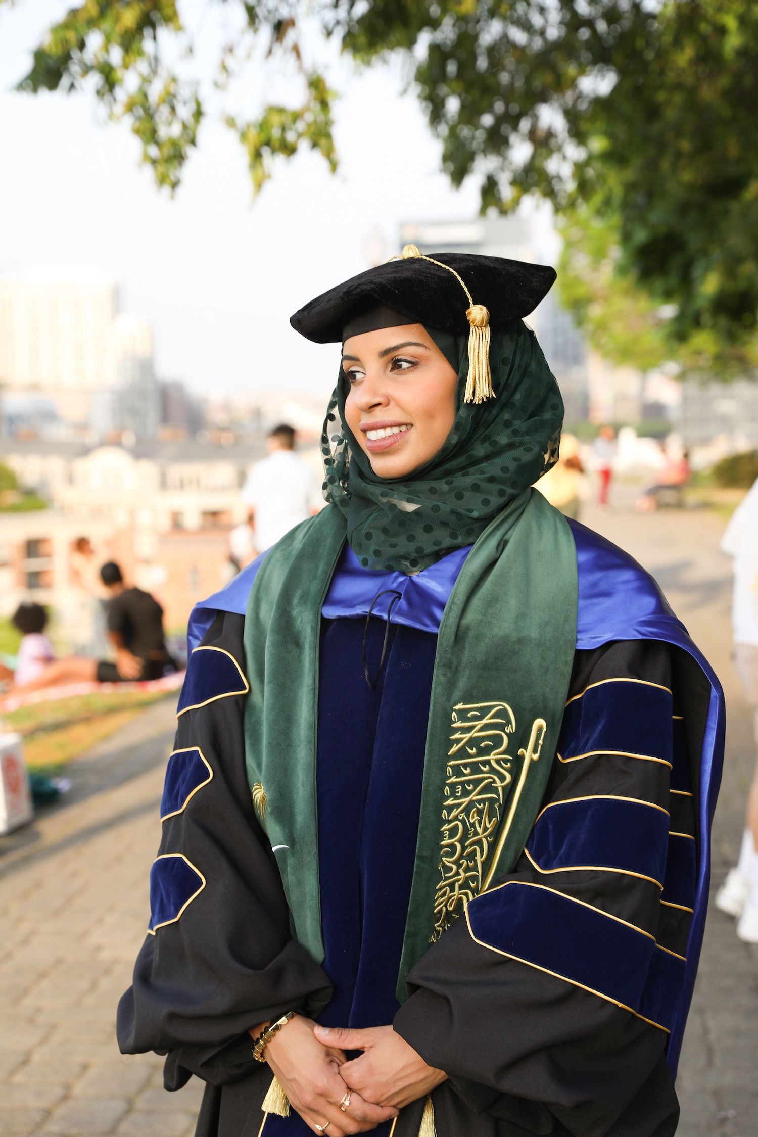 Graduate wearing navy blue and green academic regalia poses during commencement ceremony outdoors.