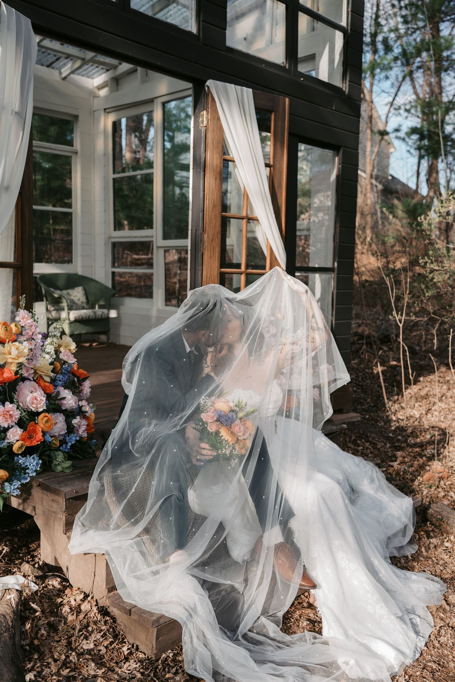 Bride and groom share a romantic kiss under a flowing veil on steps of a rustic glass cabin with colorful flowers.