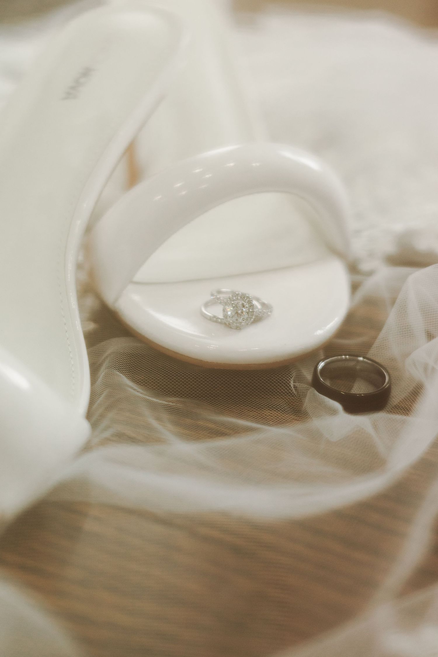 Wedding rings displayed on a white ceramic dish with soft tulle fabric, featuring a diamond engagement ring and dark band.