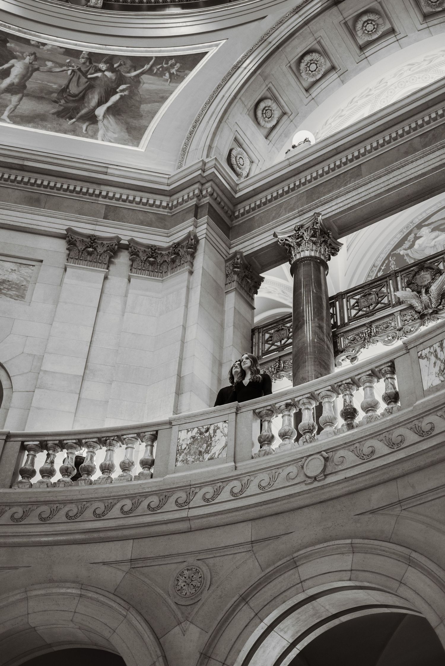 Black and white photo of two people standing on ornate balcony inside grand classical building with columns and painted ceiling.