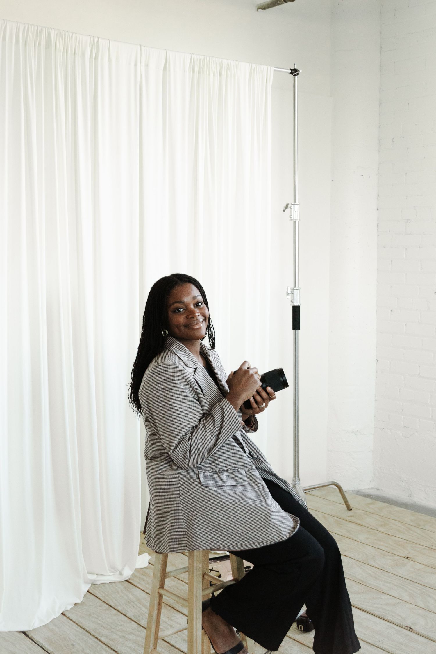 Professional photographer in a gray blazer holding camera equipment while sitting on wooden stool in studio setting.