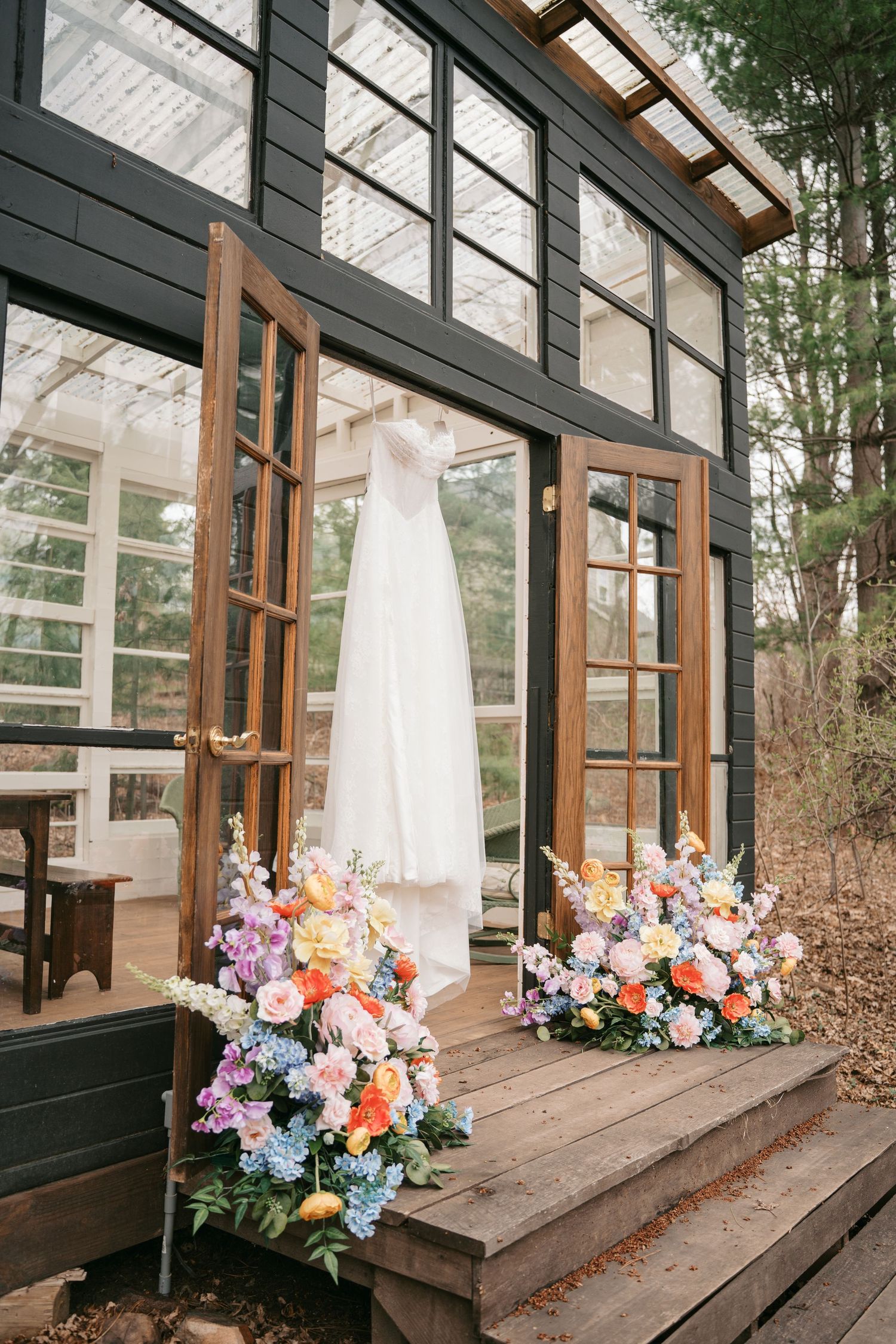 Wedding dress hanging in open French doors of black greenhouse with colorful floral arrangements on wooden steps.