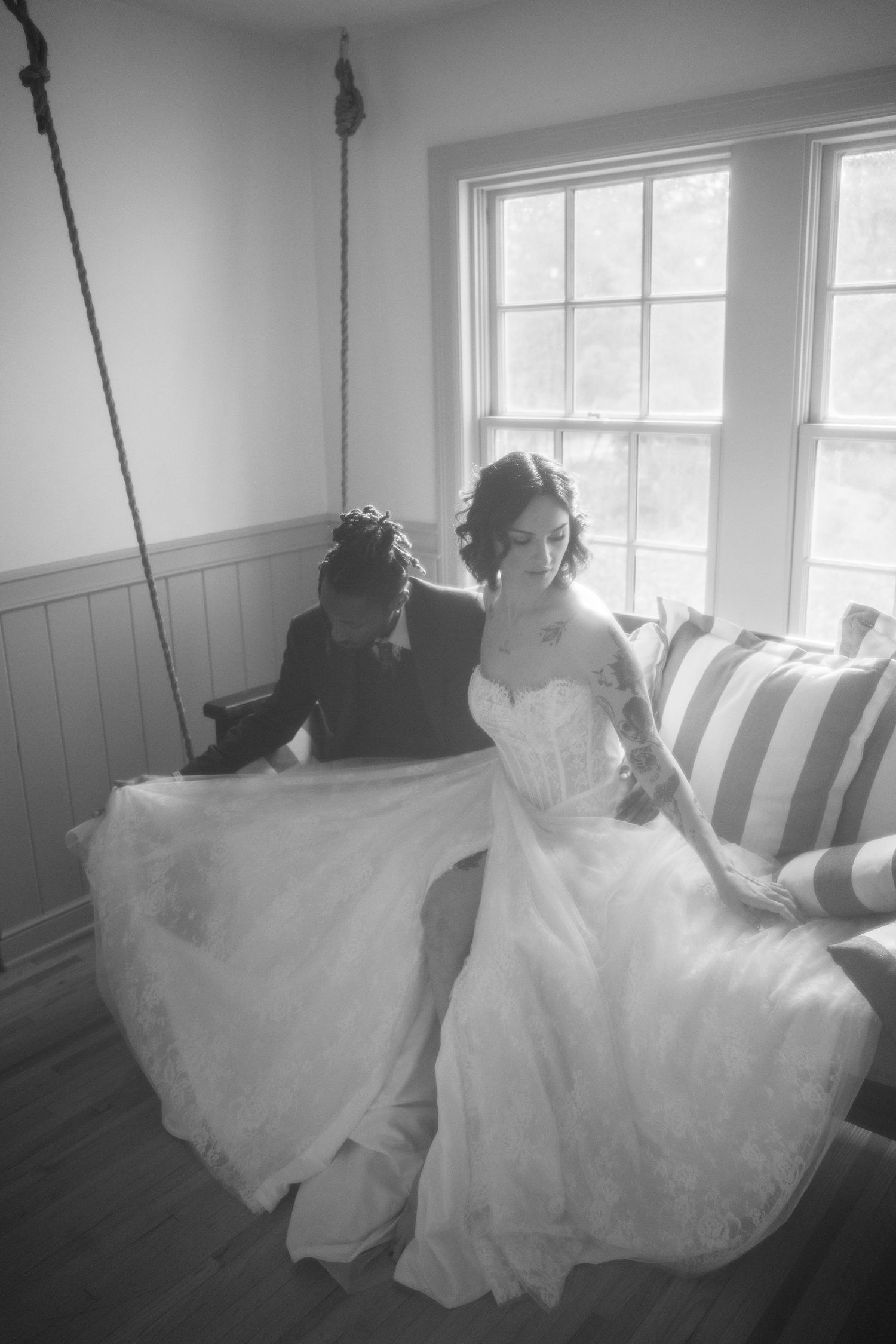 Black and white photo of a bride in a white gown being helped by a bridesmaid near a sunlit window.