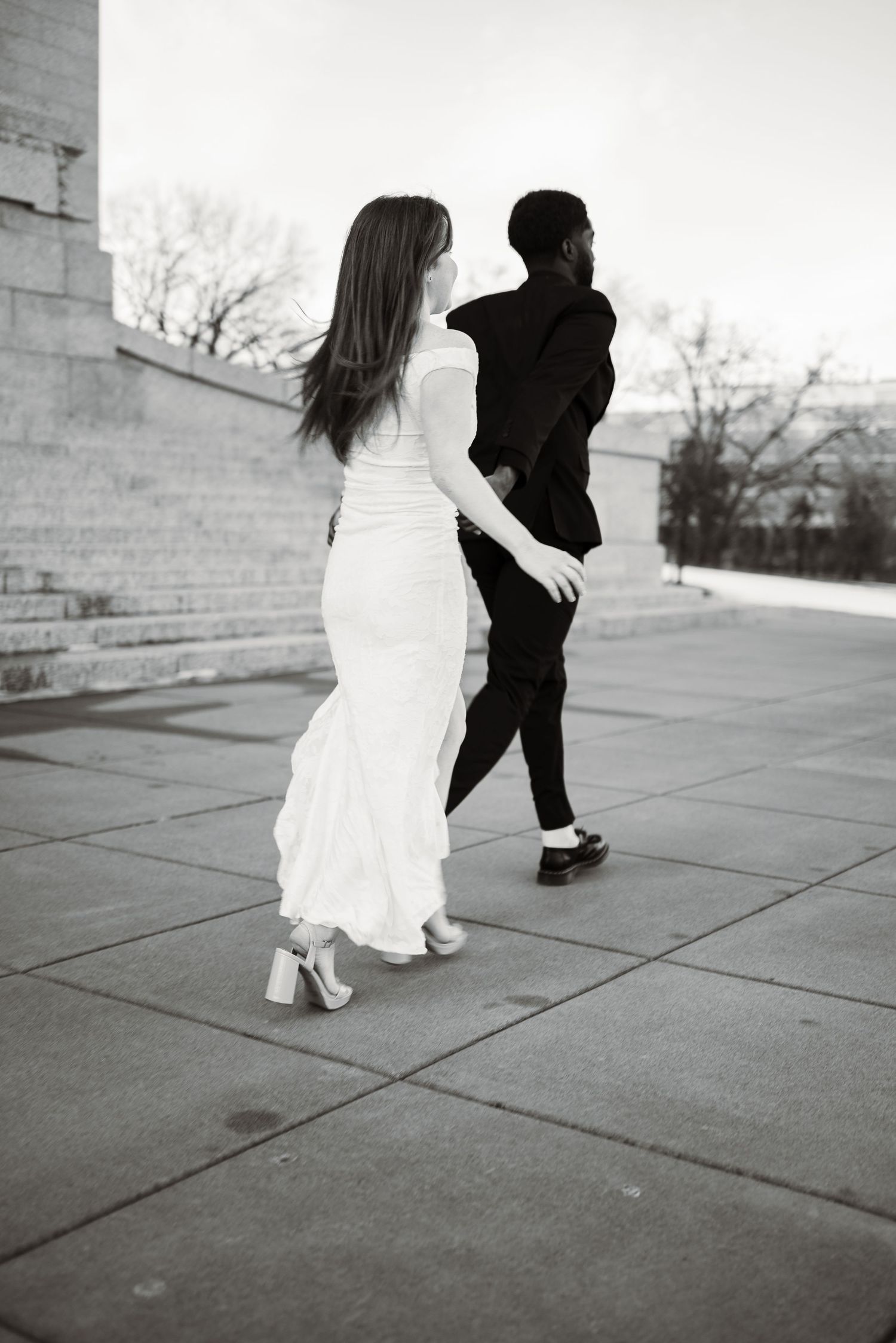 A bride in a white gown and groom in a black suit walk hand in hand on a stone plaza in a black and white photo.