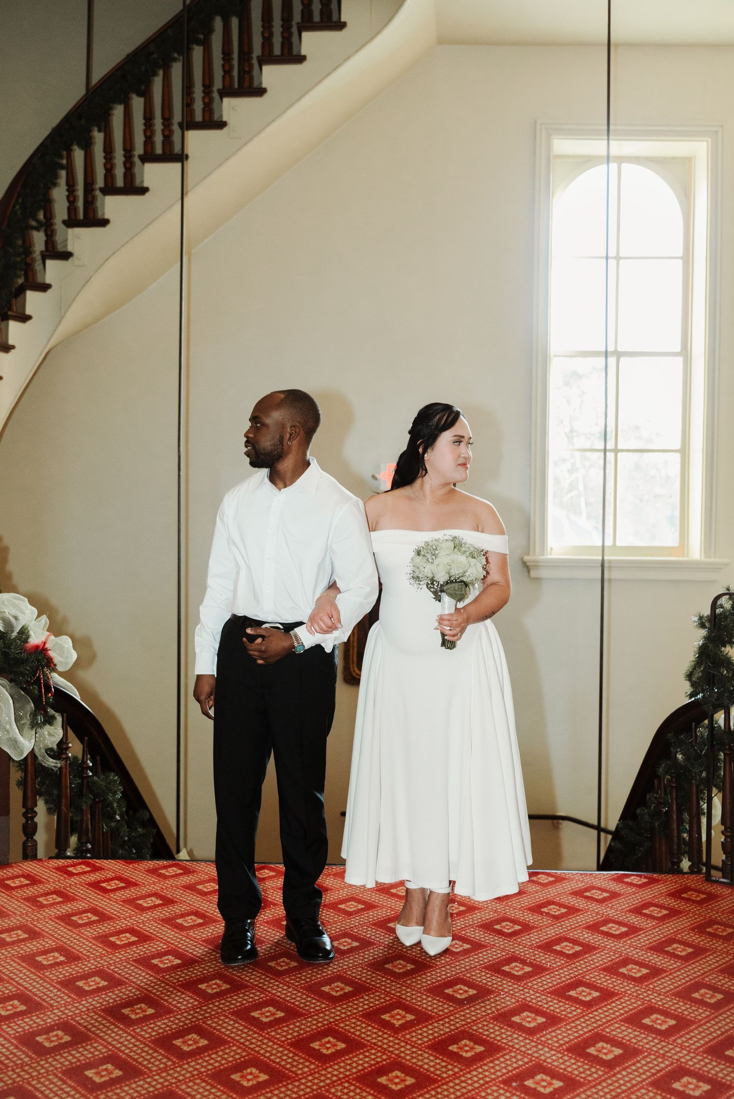 Bride and groom stand together in elegant venue with grand staircase, bride holding white bouquet in off-shoulder tea-length gown.