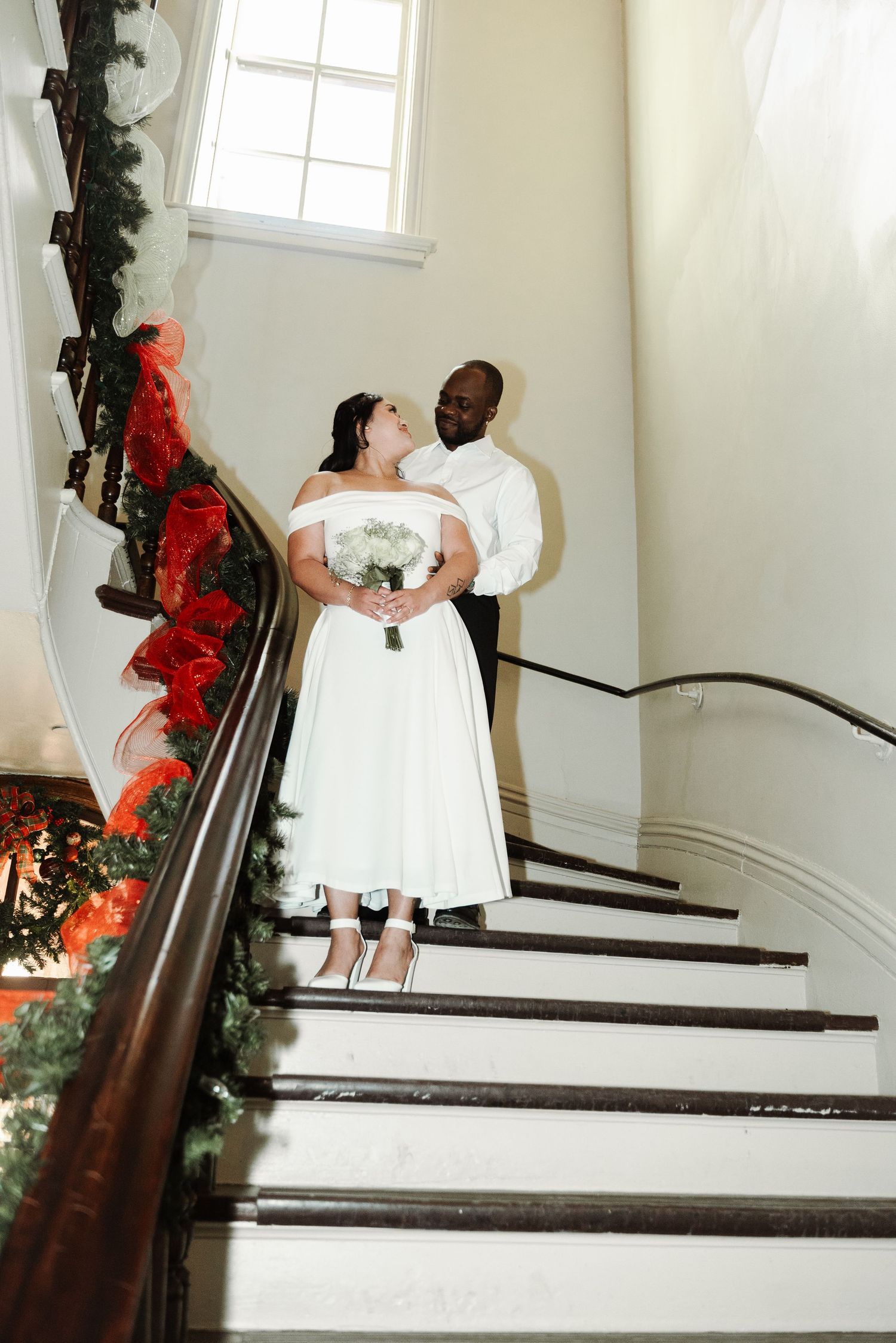 Bride and groom pose on decorated staircase, bride in white off-shoulder gown holding bouquet, holiday greenery backdrop.