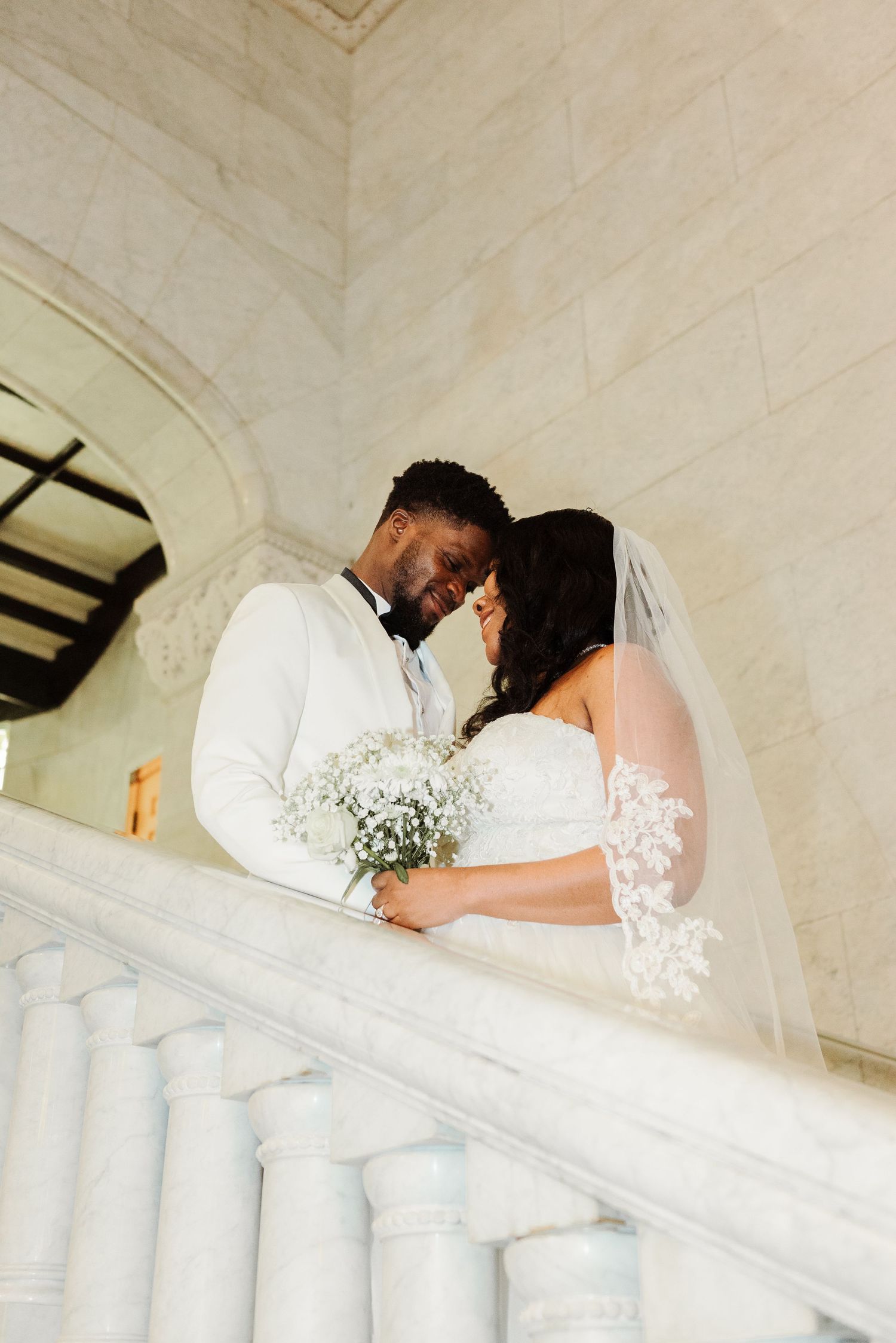 Newlywed couple in white wedding attire sharing a romantic moment on marble staircase with baby's breath bouquet.