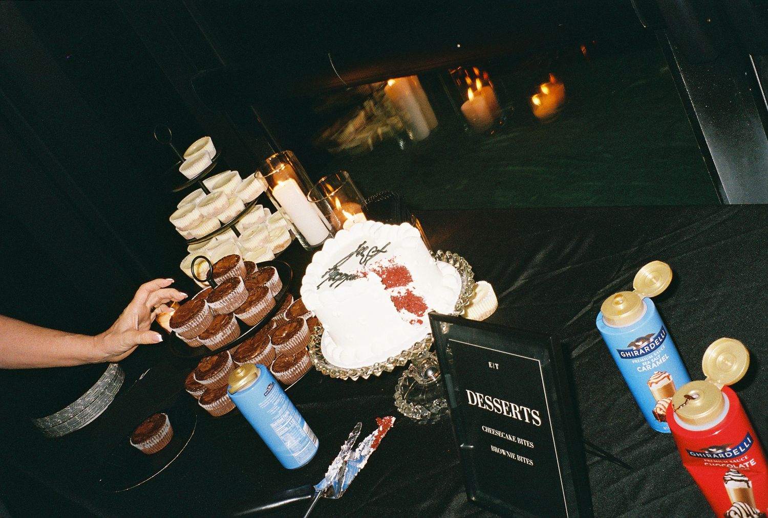 Dessert table with layered cake, chocolate cups, and colorful canisters at a dark event venue.