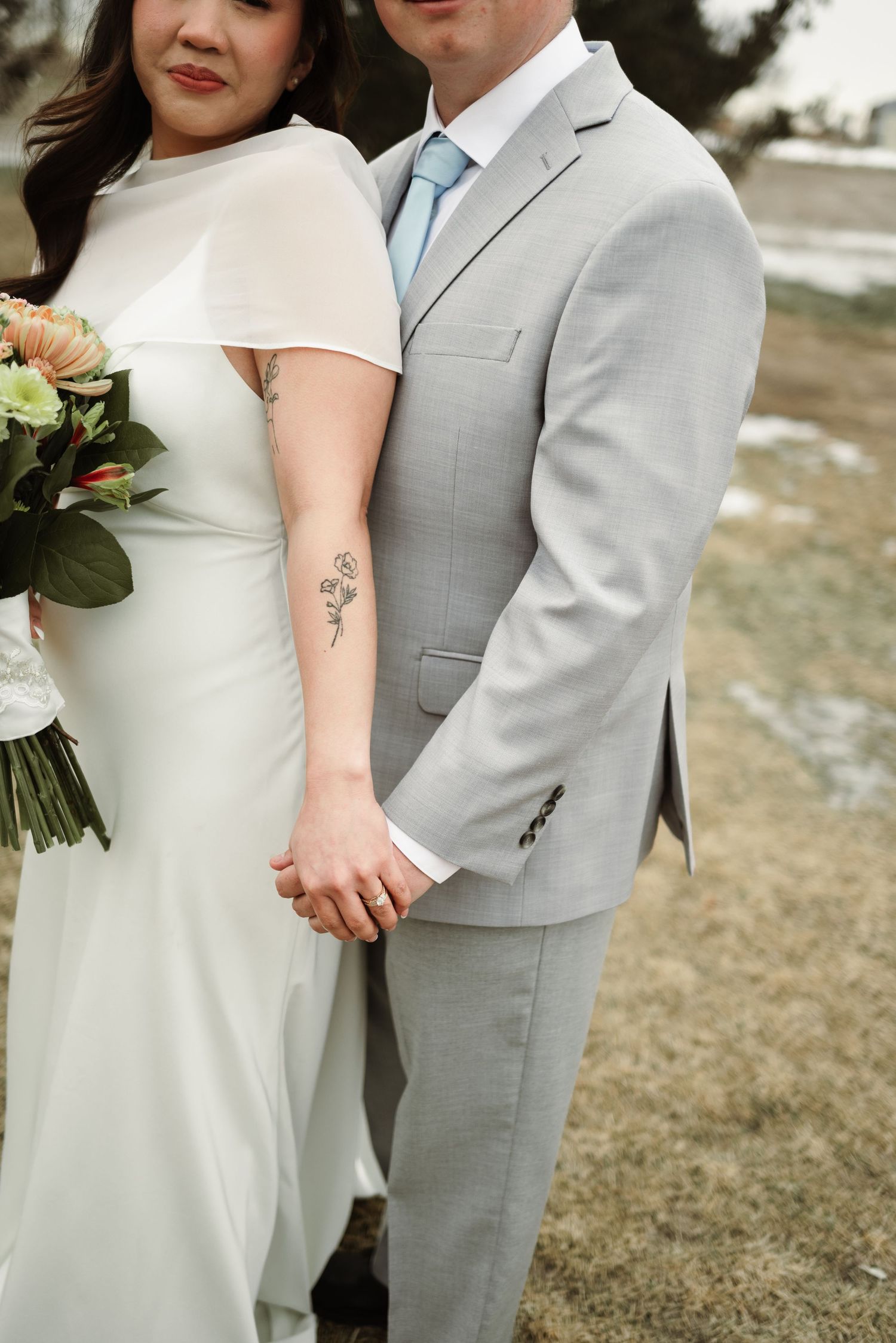 Bride in white cape gown with floral tattoo holds hands with groom in light gray suit outdoors.