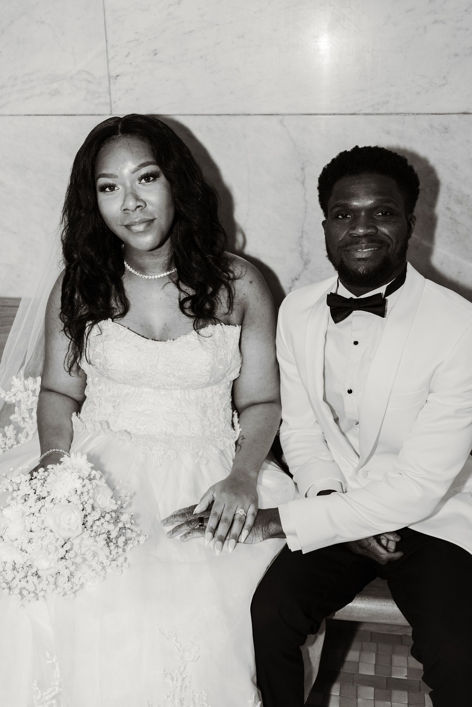 Black and white wedding portrait of a bride in lace gown and groom in white tuxedo seated together.