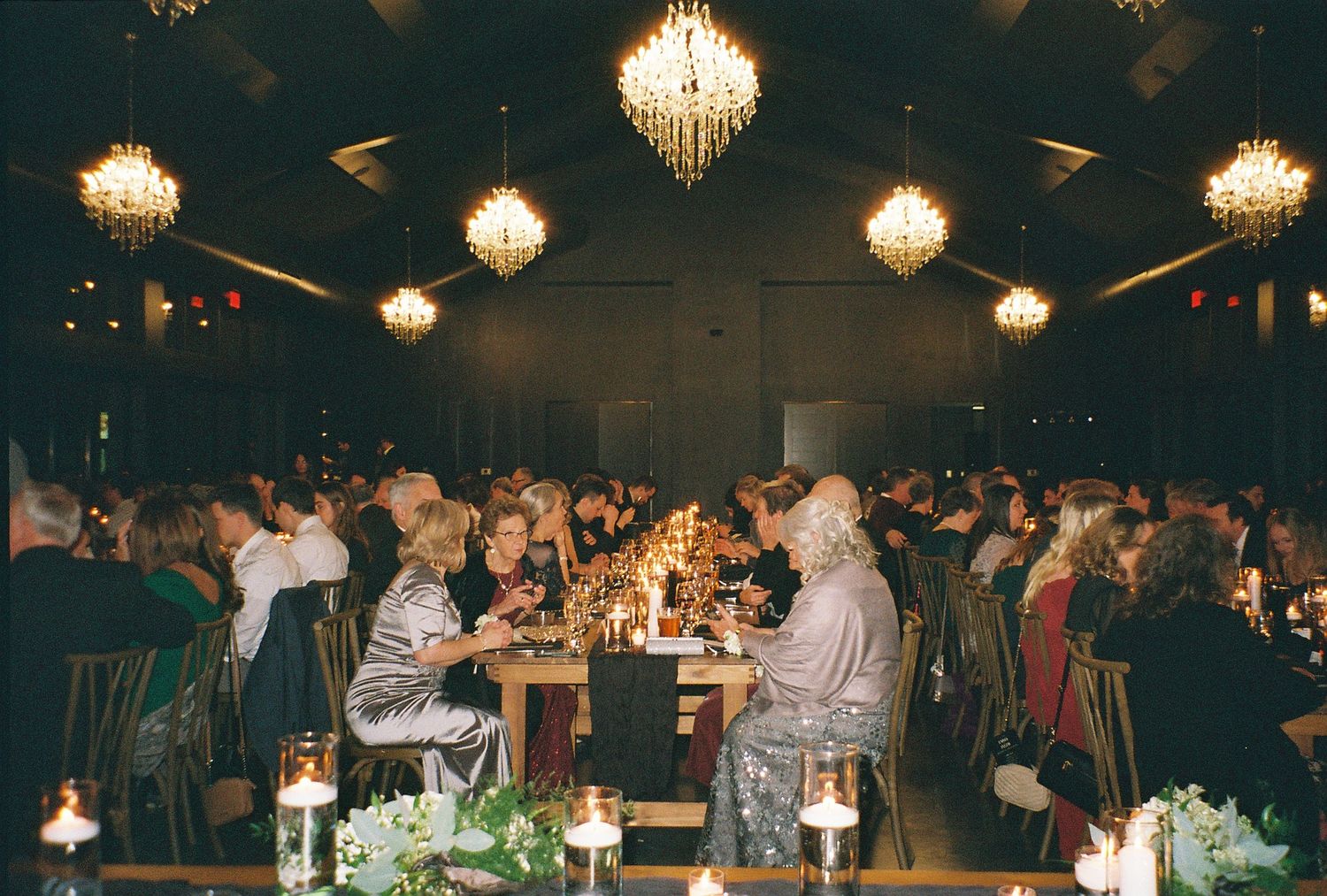 Elegant gala dinner event with guests seated at candlelit tables beneath golden chandeliers in a dimly lit banquet hall.