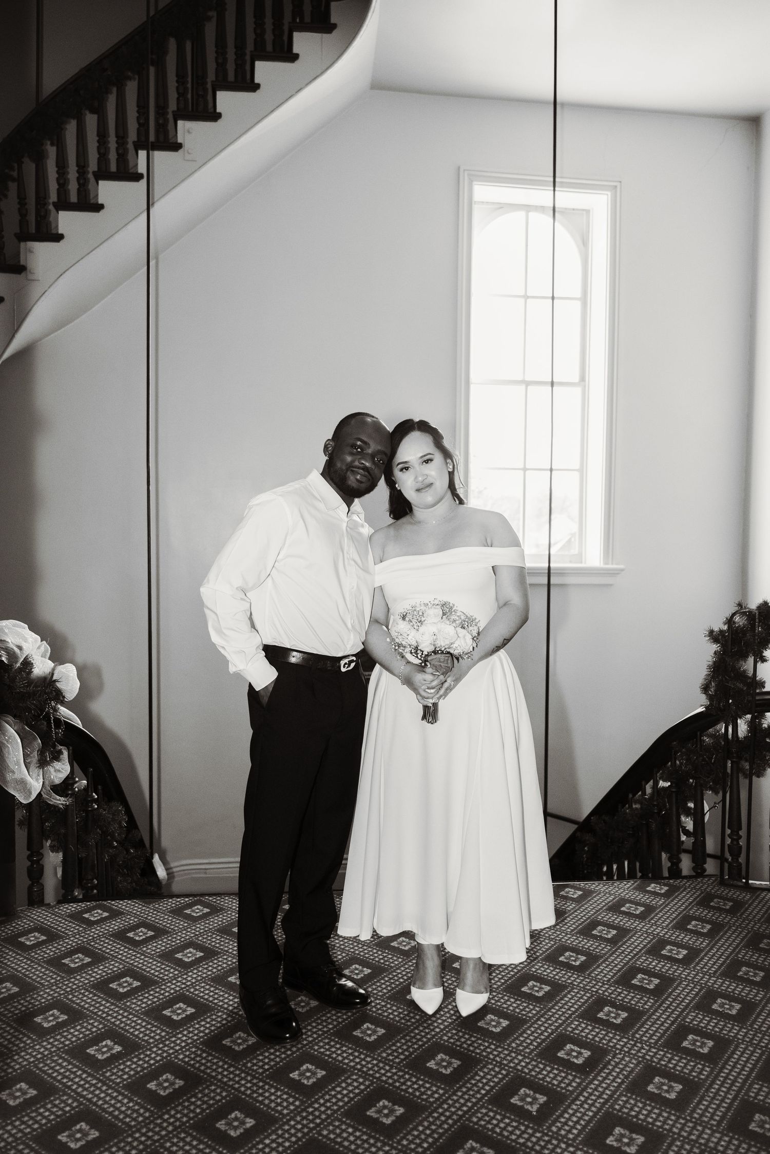 Black and white wedding photo of a couple posing elegantly on a staircase, bride in tea-length gown holding bouquet.