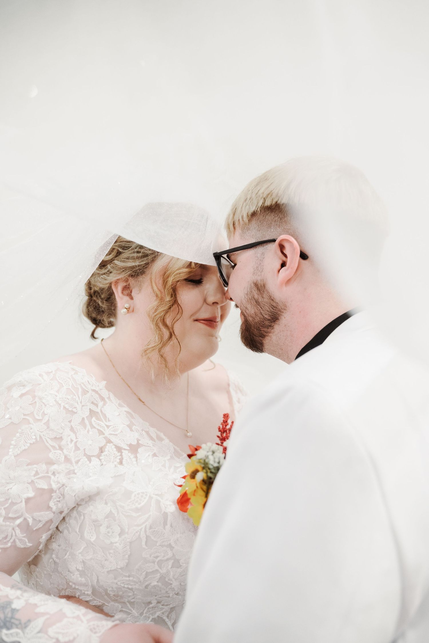 A bride and groom share an intimate moment, nearly kissing, dressed in white wedding attire with a floral bouquet.