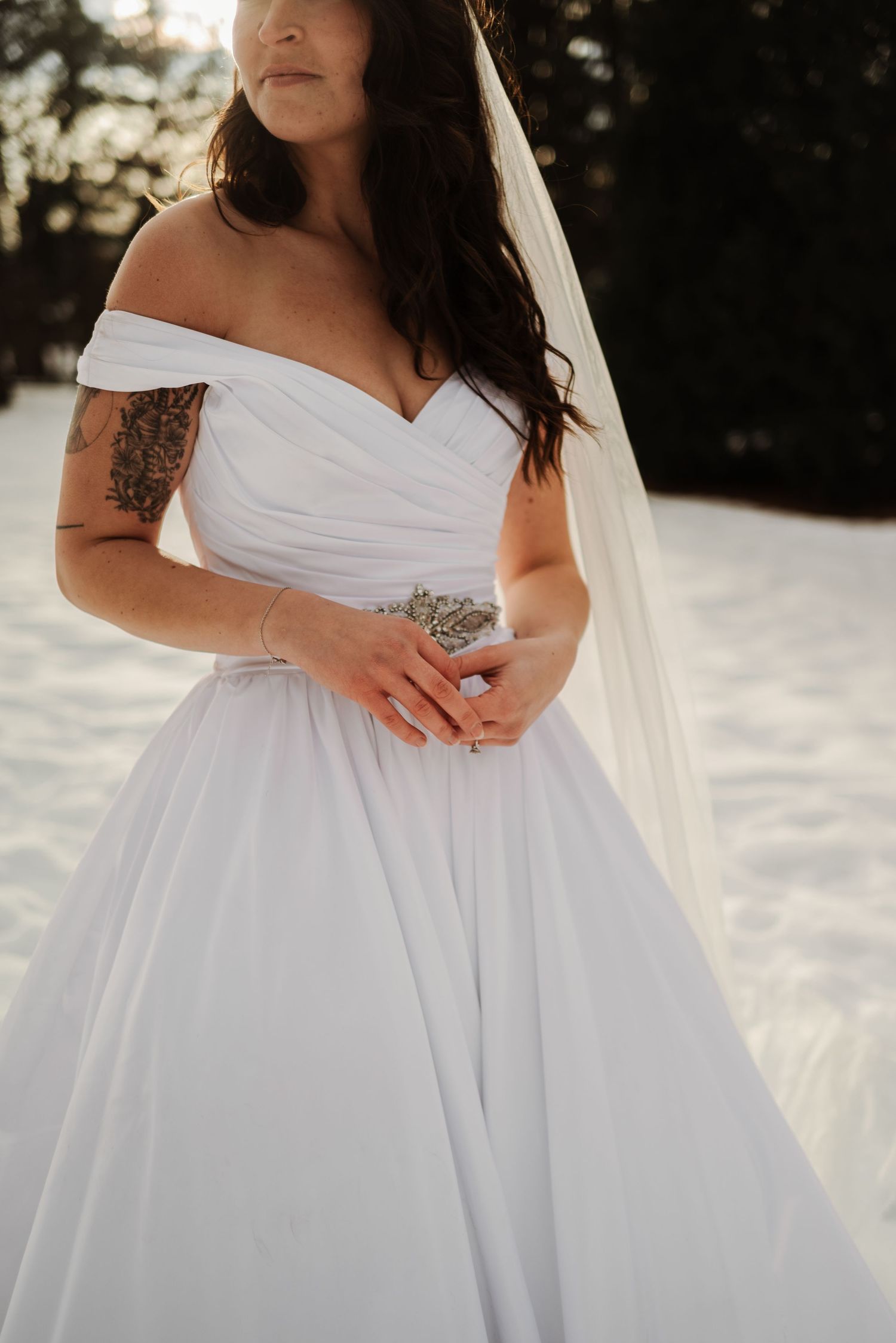 Bride in off-shoulder white ball gown with veil and arm tattoo, posing in snowy winter outdoor setting.