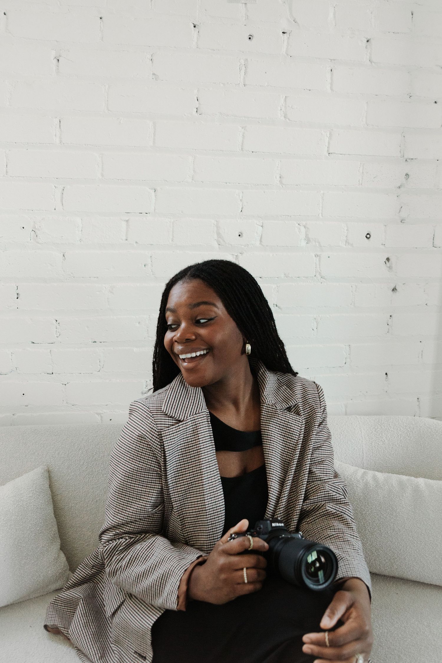 Photographer with camera smiles while sitting on white couch against brick wall.
