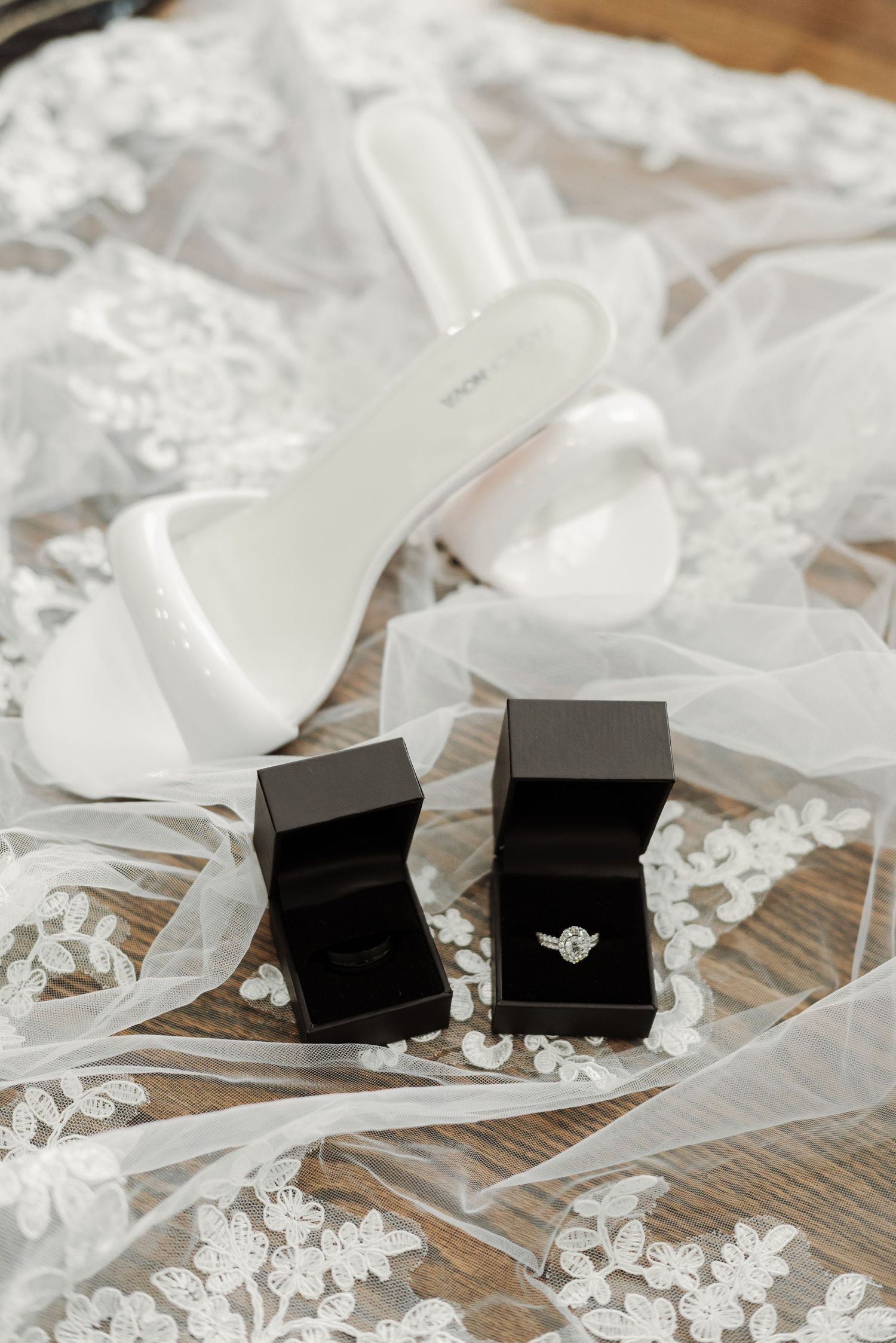 Wedding accessories including white heels and black ring boxes arranged on delicate lace fabric.