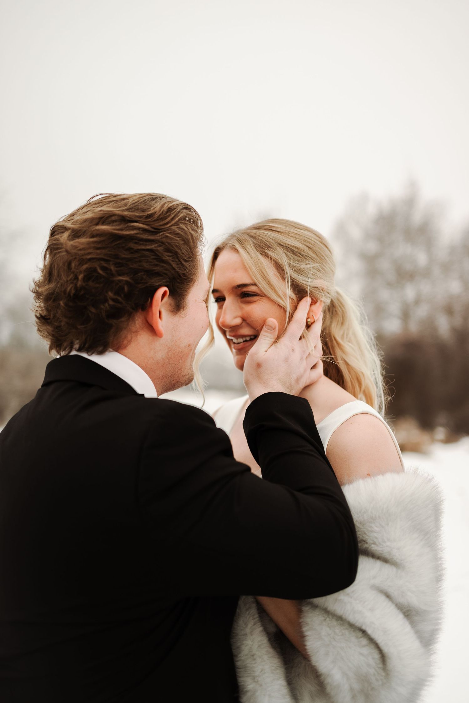 Couple sharing romantic winter moment, groom tenderly cupping bride's smiling face outdoors in snowy landscape.