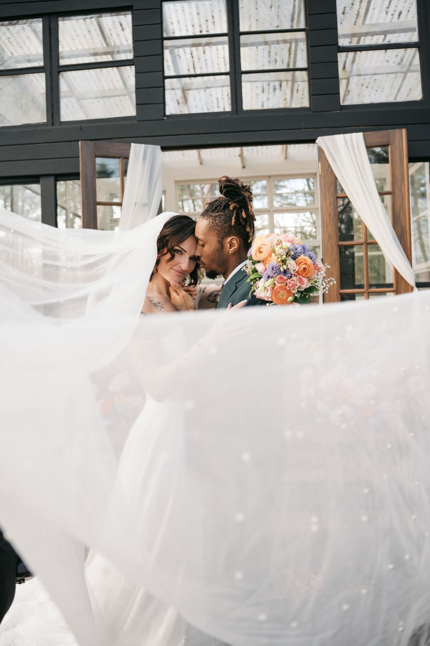 Bride and groom share romantic moment under white draped canopy with colorful bouquet, veil flowing in foreground.