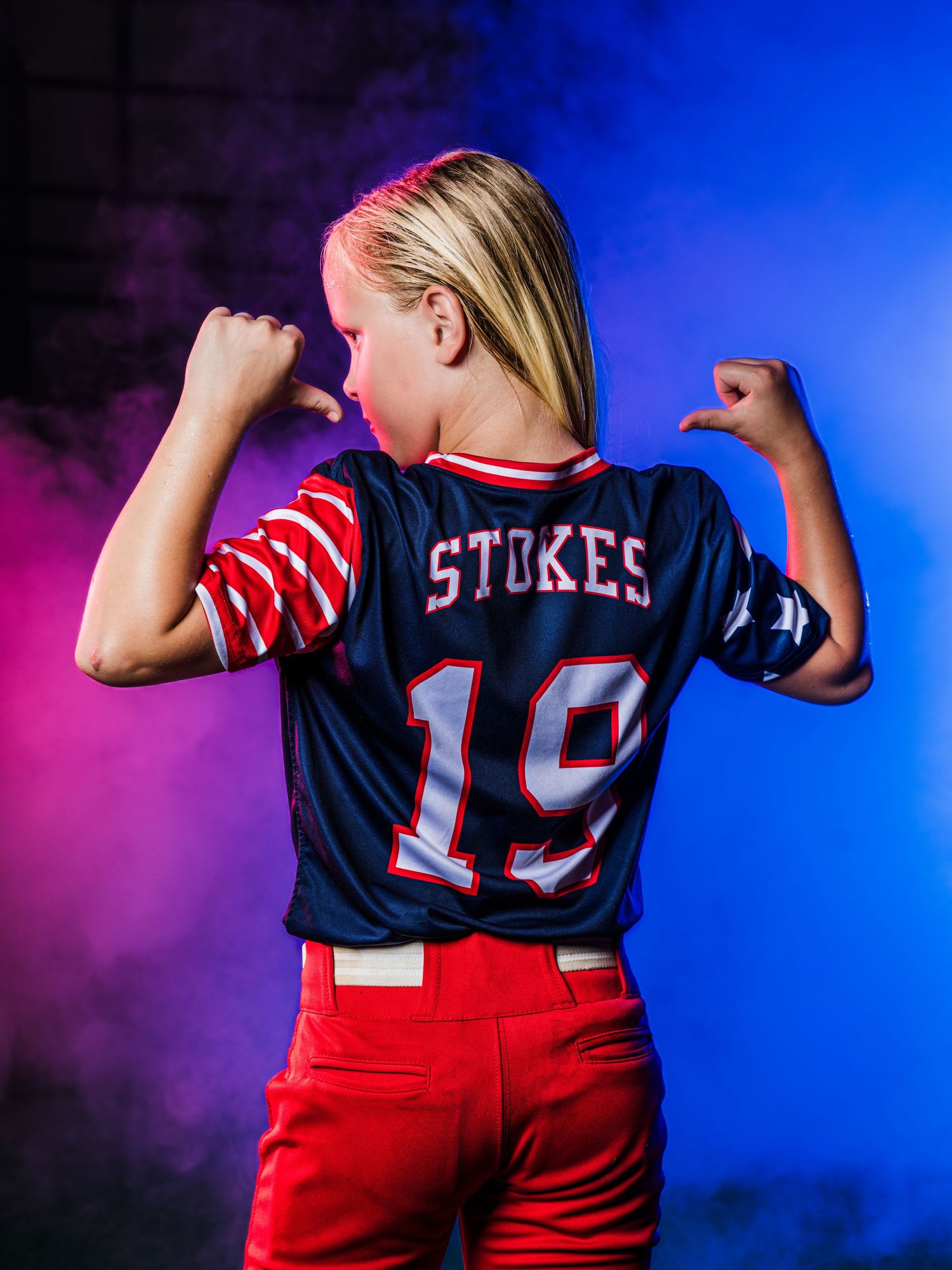 Young athlete poses in navy Stokes #19 baseball jersey with red pants against colorful smoky background.