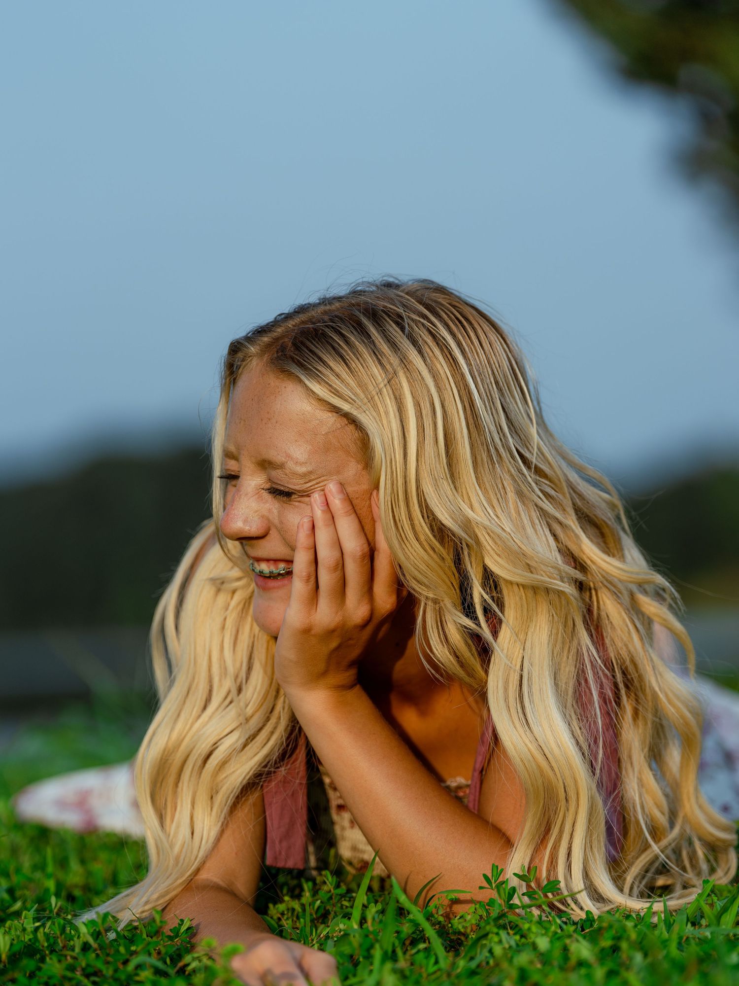 Young blonde girl lying on grass, smiling and resting her chin on her hands on a sunny day.