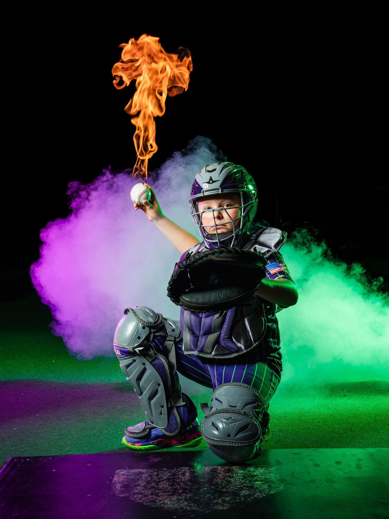 Young baseball catcher in full gear holds flaming ball amid colorful green and purple smoke on dark background.