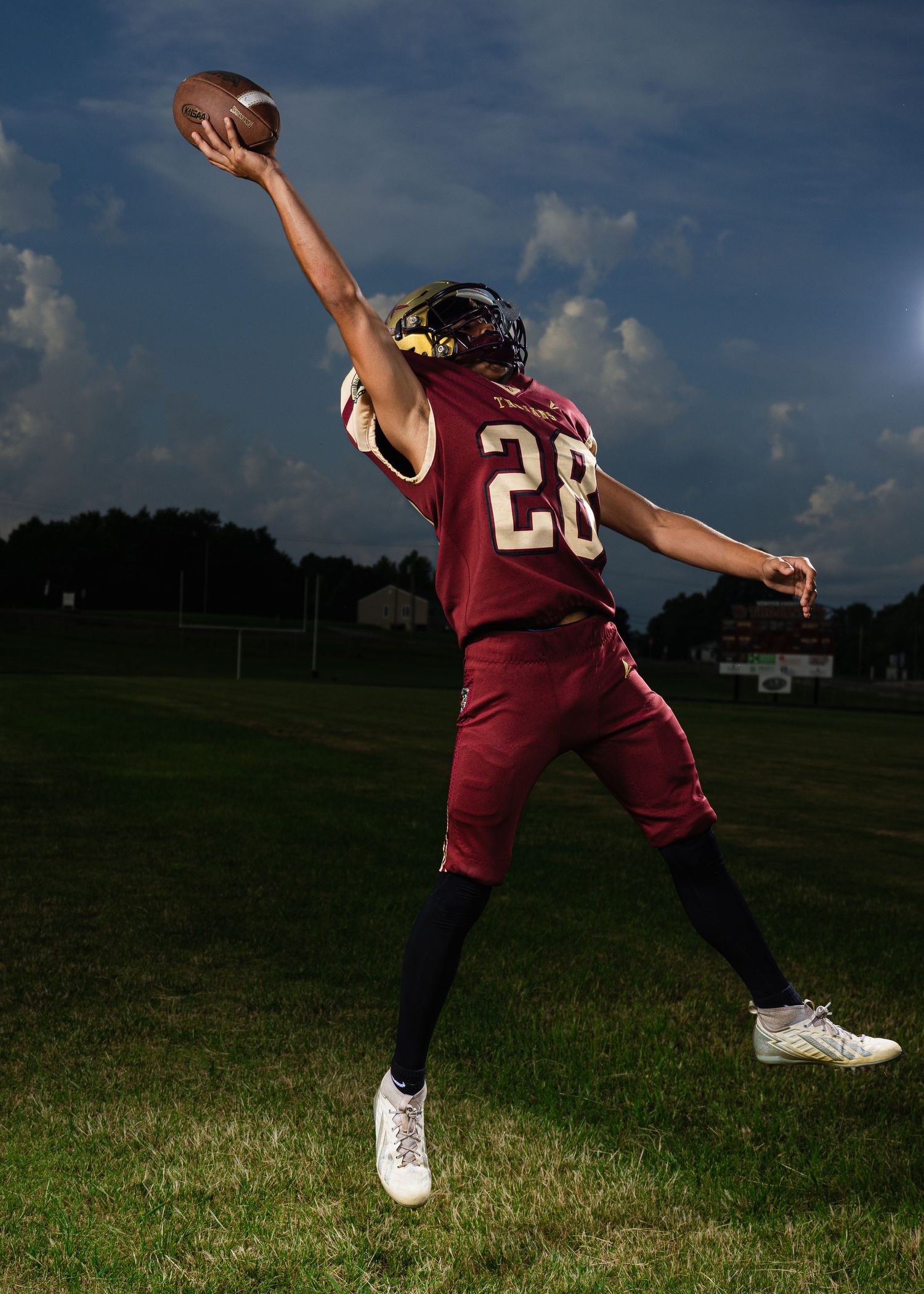 Football player #28 in maroon uniform leaps to catch a ball one-handed against a dramatic cloudy sky on a green field.