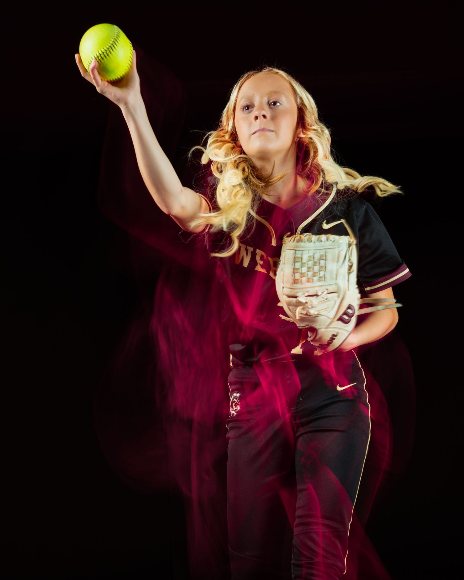 Young female softball pitcher in dark uniform winds up to throw a neon yellow ball against a dramatic dark background.