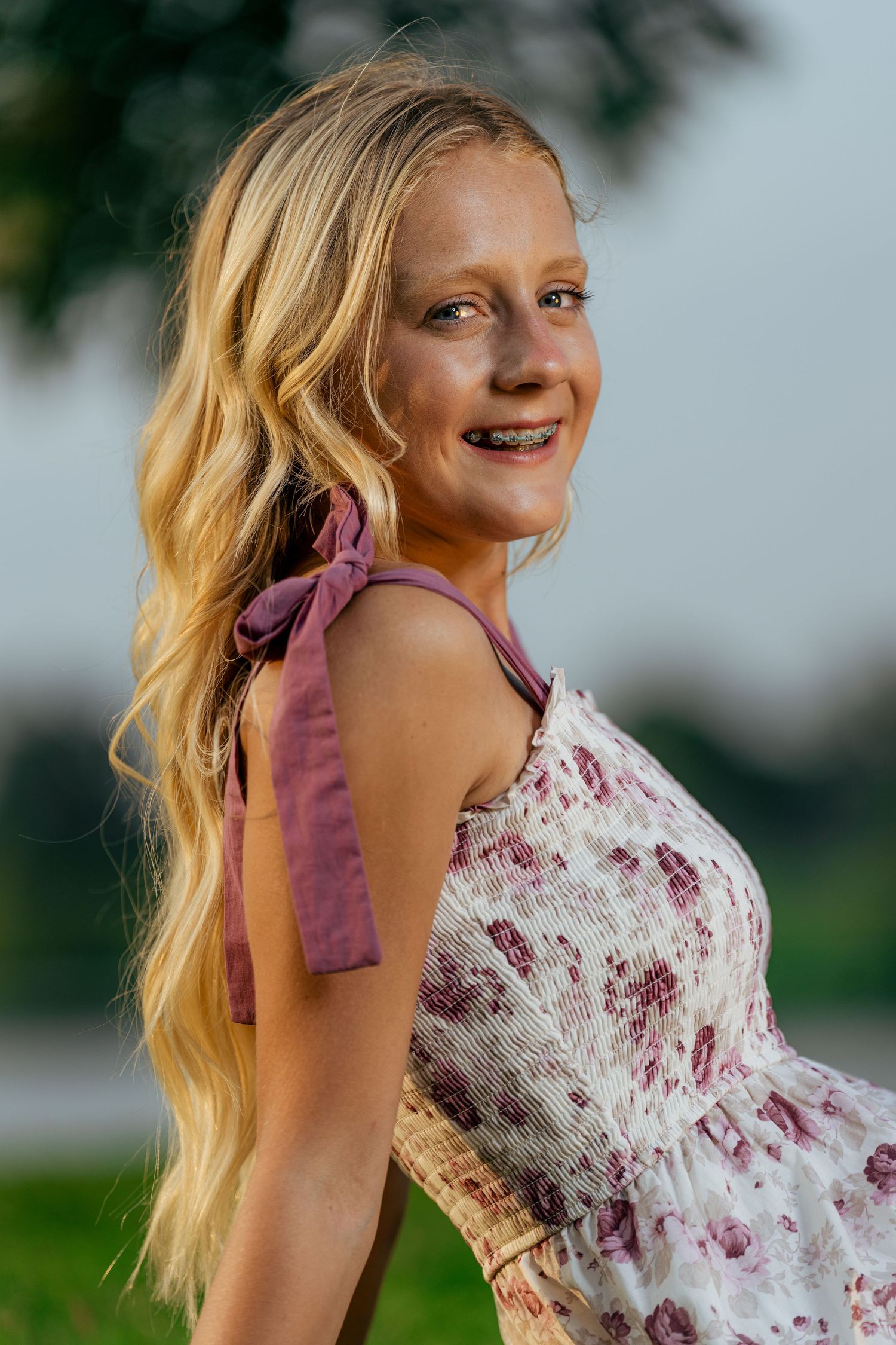 Smiling blonde teenage girl with braces wearing a floral sundress outdoors in soft natural light.