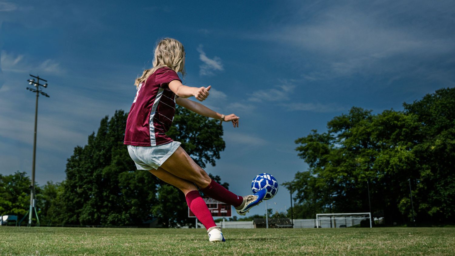 Female soccer player in maroon uniform leaping mid-air to kick a soccer ball on a green field under blue sky.