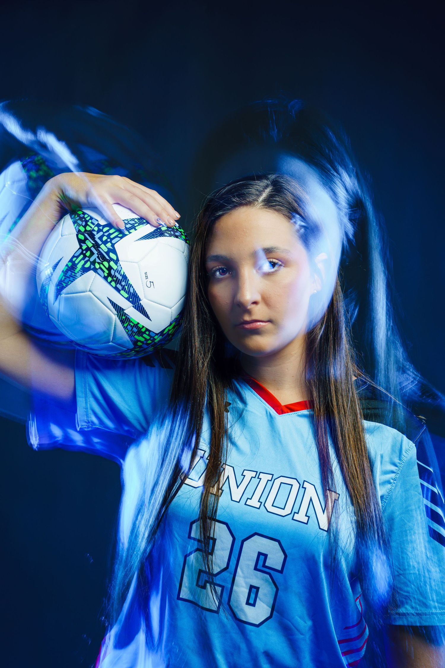 Young girl in Union #26 soccer jersey holding a ball on her shoulder, posed against a dark blue background with light streaks.