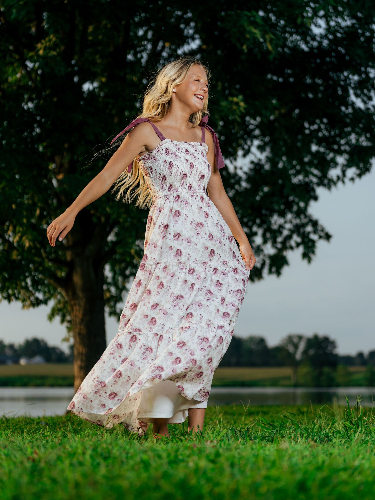 Young girl twirling in a floral maxi dress on green grass near a tree and lake.