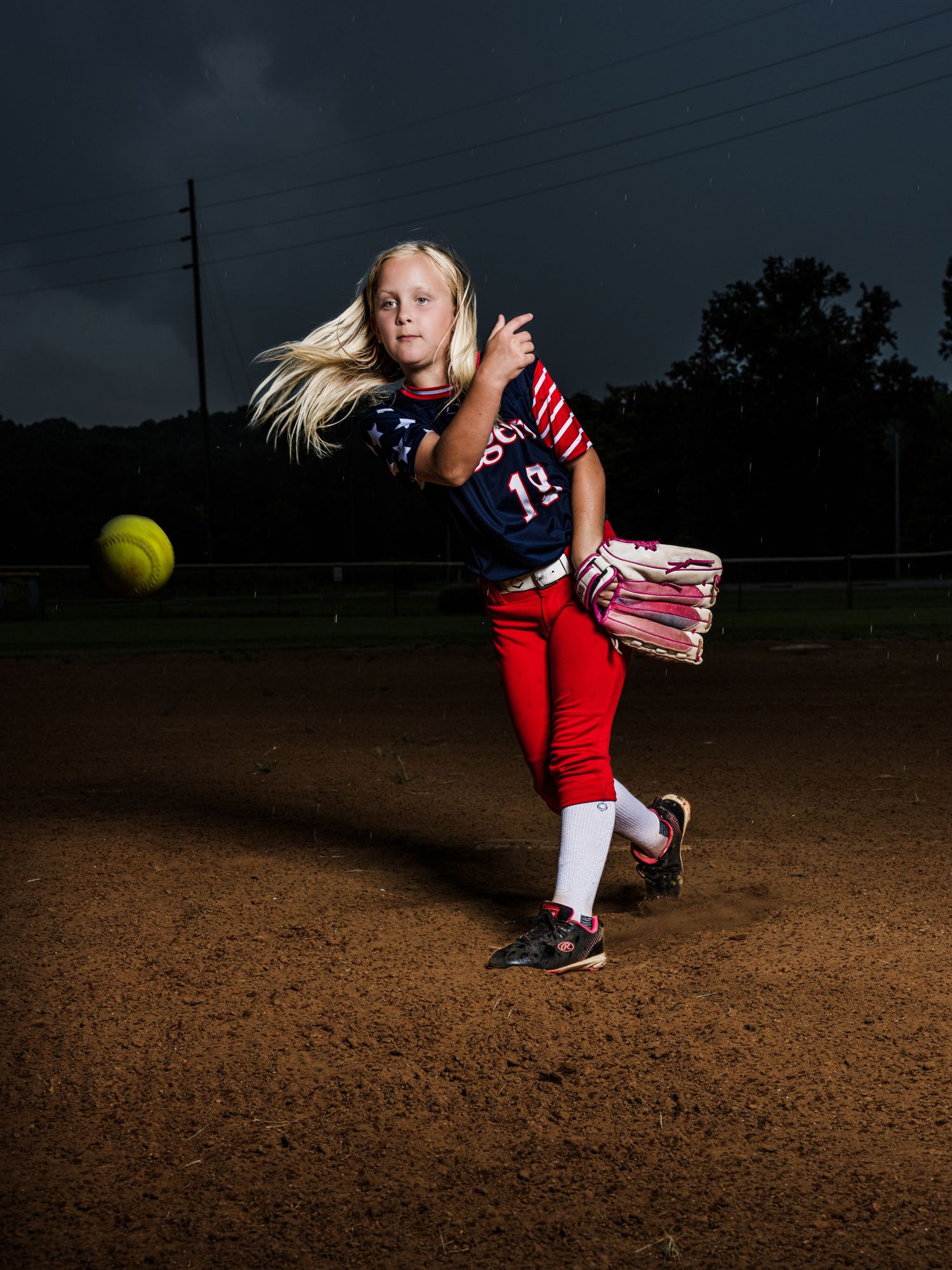 Young blonde girl in navy and red softball uniform pitching a yellow ball on a dirt mound under dramatic stormy sky.