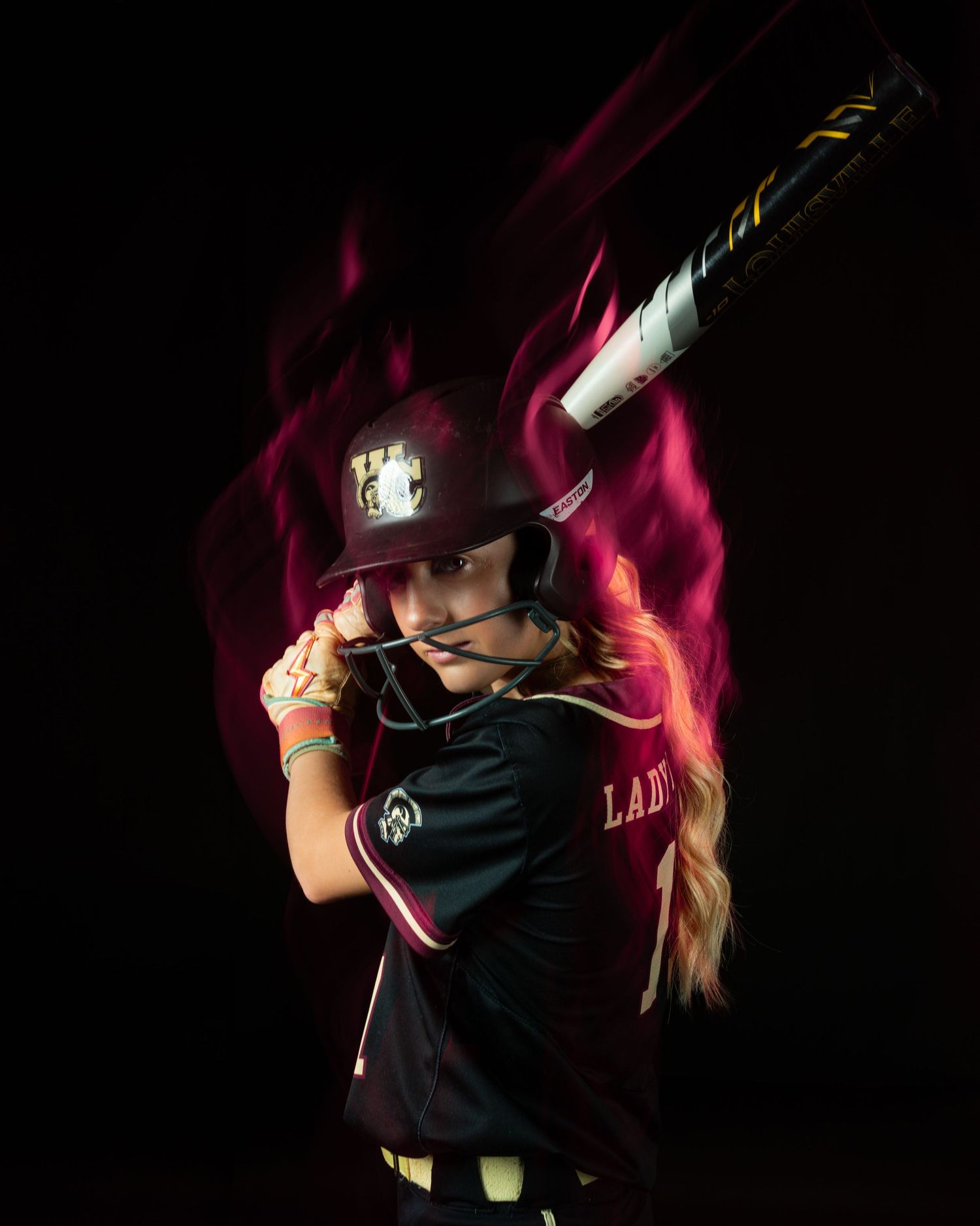 Female softball player in dark uniform poses with bat over shoulder, dramatic pink flame effect on black background.
