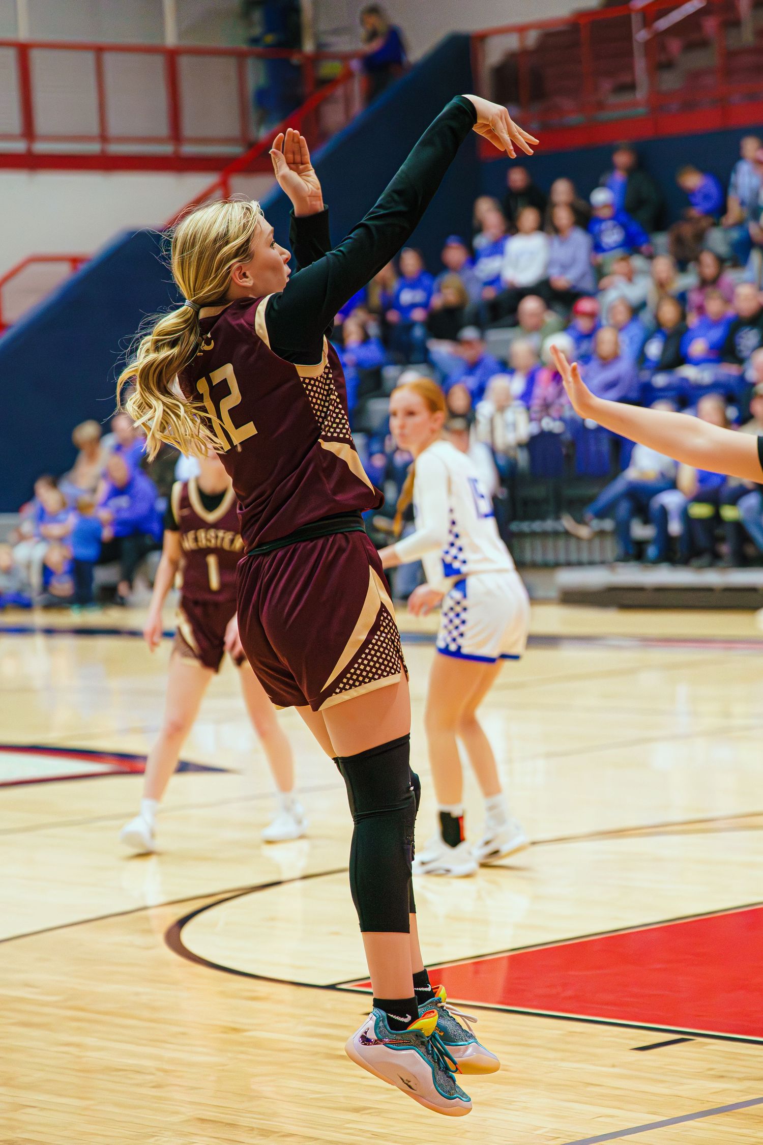 Female basketball player in maroon jersey #12 shoots the ball during a competitive girls high school basketball game.