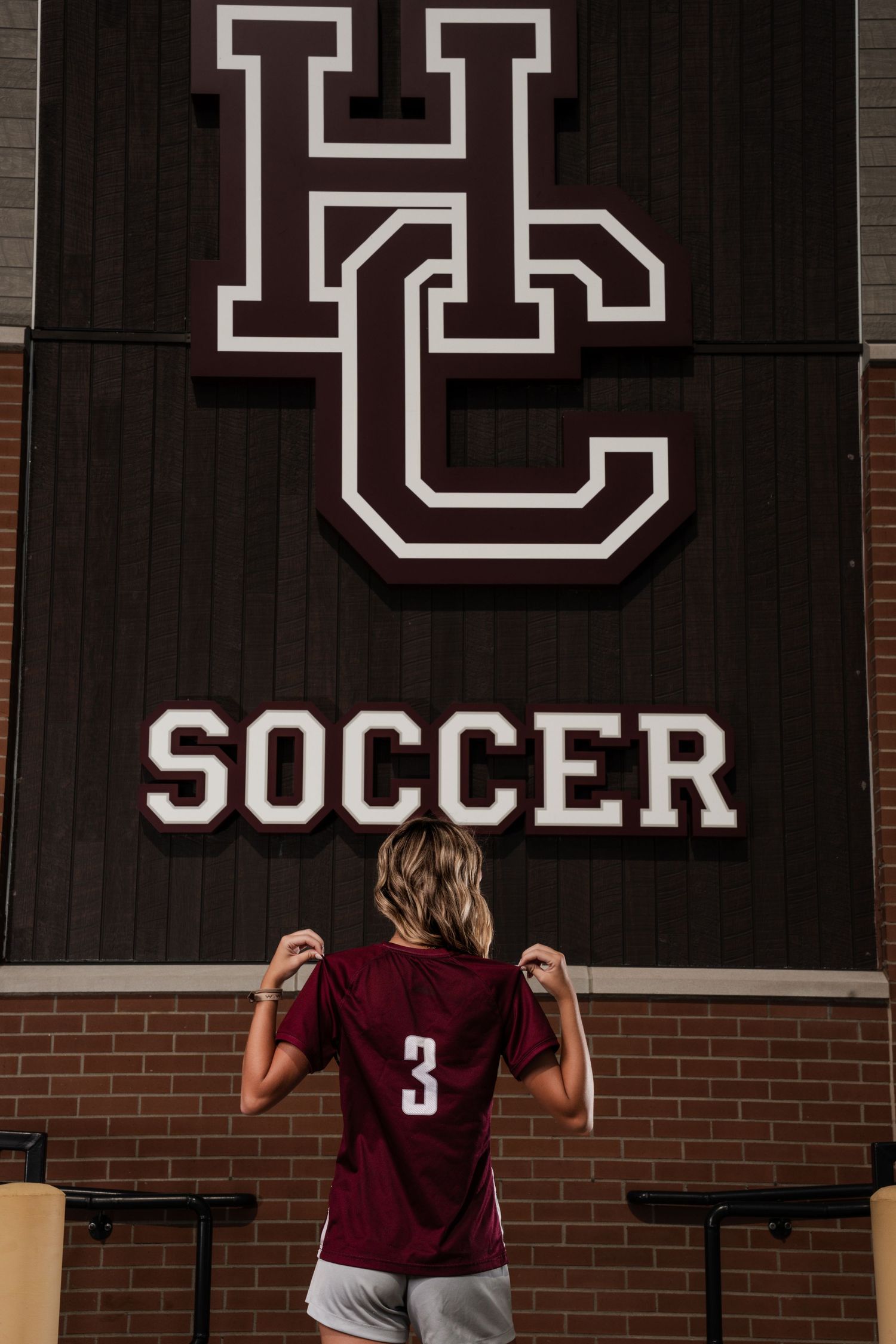 Female soccer player #3 stands before HC Soccer sign in maroon jersey, showing team spirit at high school athletic facility.
