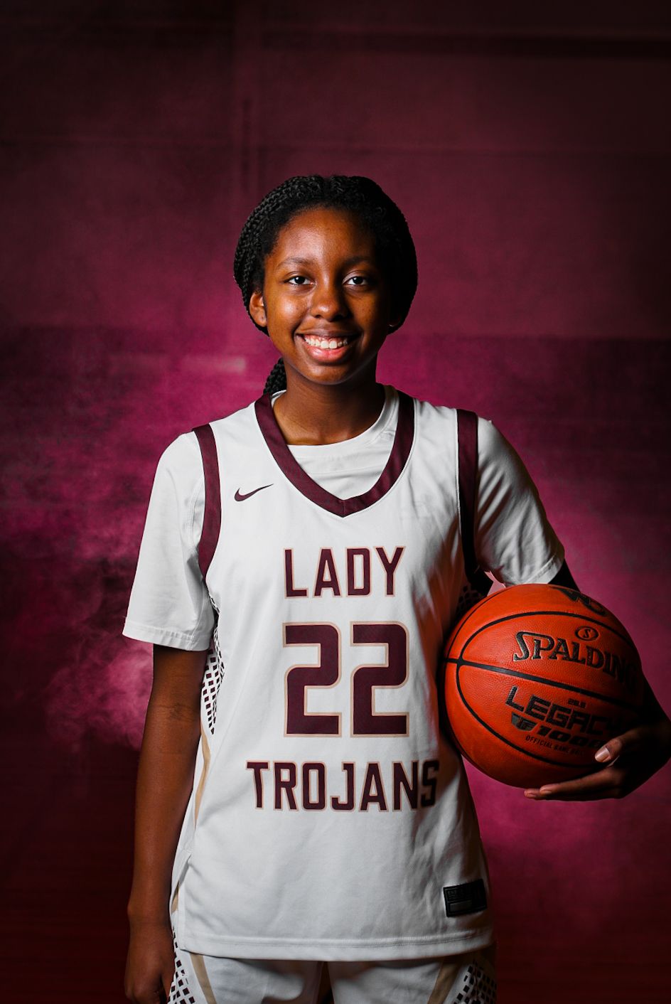 Basketball player in white Lady Trojans jersey poses with basketball against dark purple backdrop.