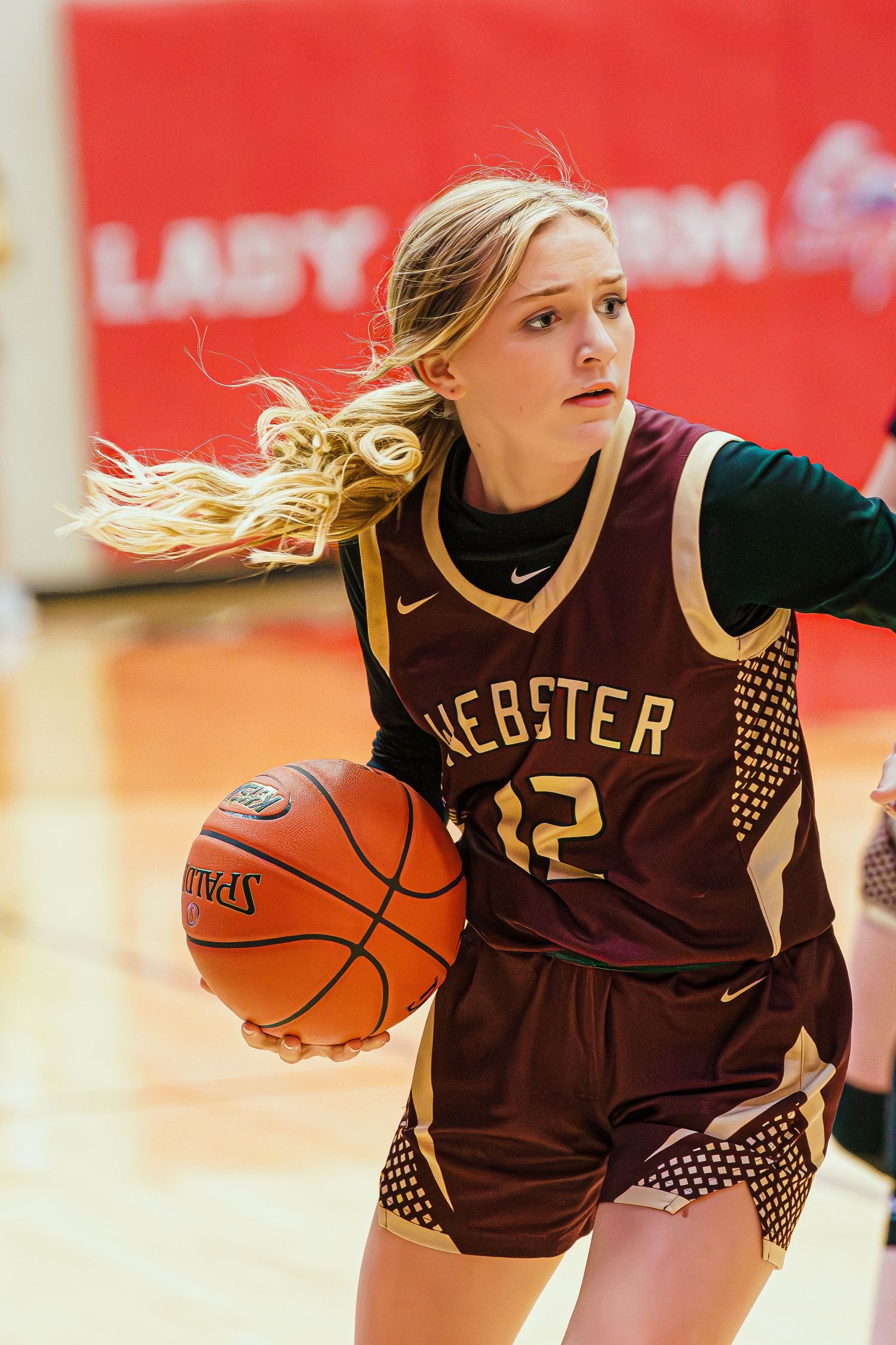A young female basketball player in a Webster #12 maroon uniform dribbles the ball during a competitive indoor game.