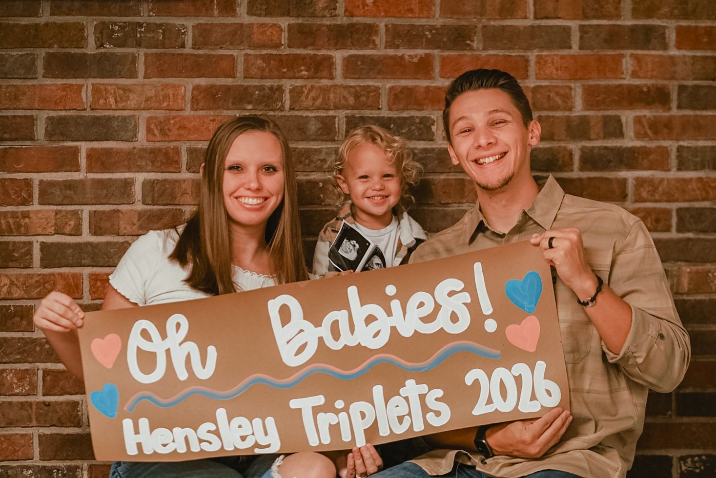 Triplets announcement photo shoot with wooden signs against brick wall.