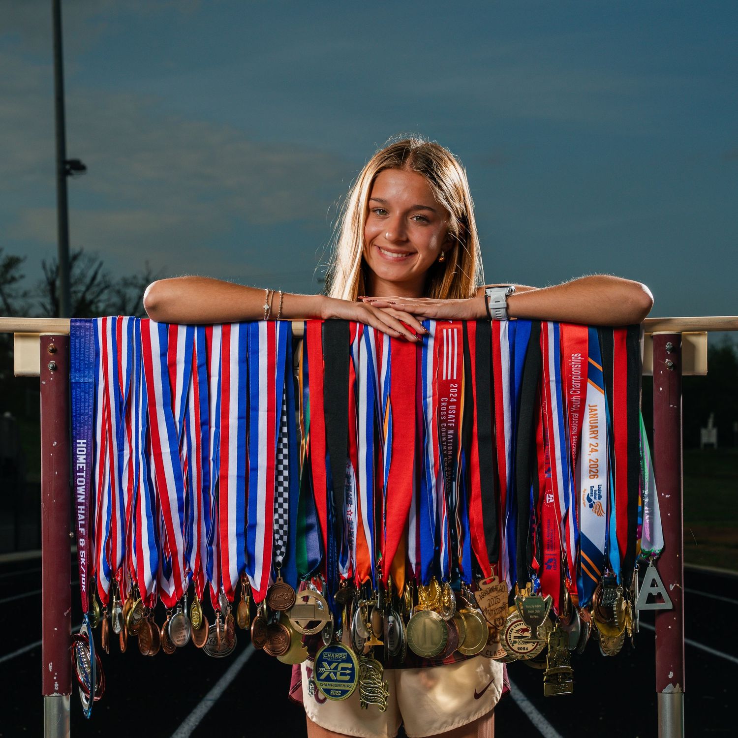 Young female athlete smiles behind a railing displaying dozens of colorful medals on a track field.