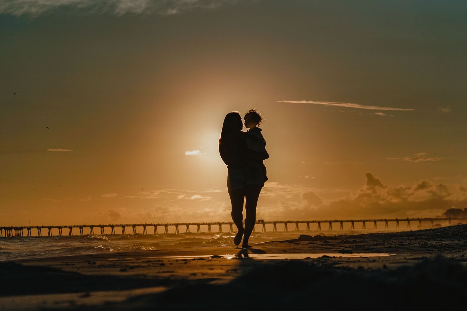 Silhouette of figure holding child against golden sunset with pier in distance.