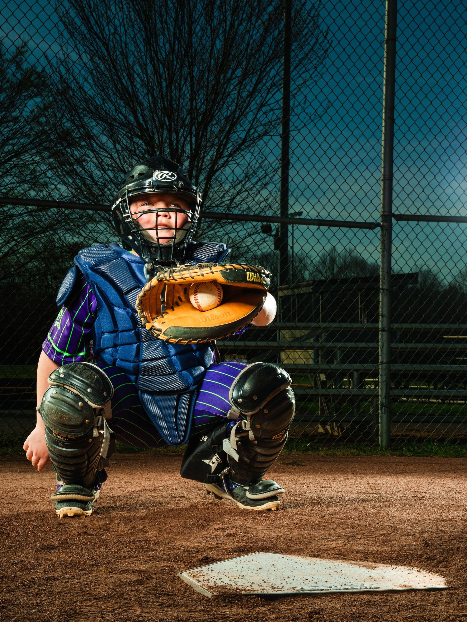 Baseball catcher in full gear crouching at home plate, catching a pitch in a leather mitt during a night game.