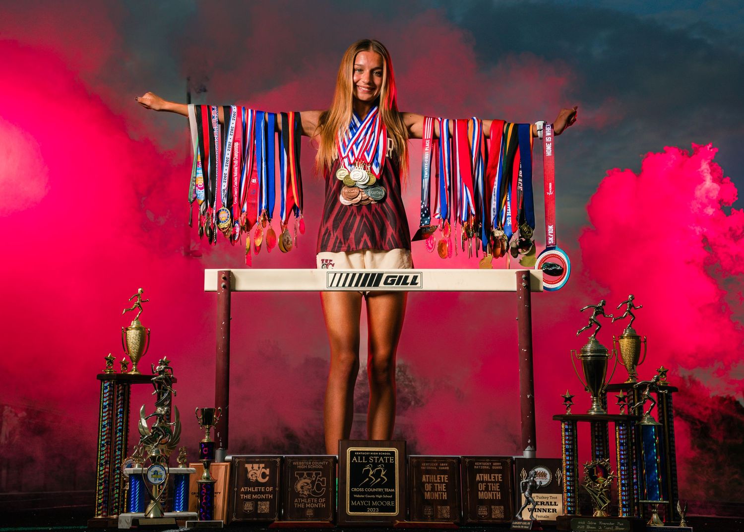 Female athlete stands triumphantly on hurdle displaying medals trophies amid pink smoke.